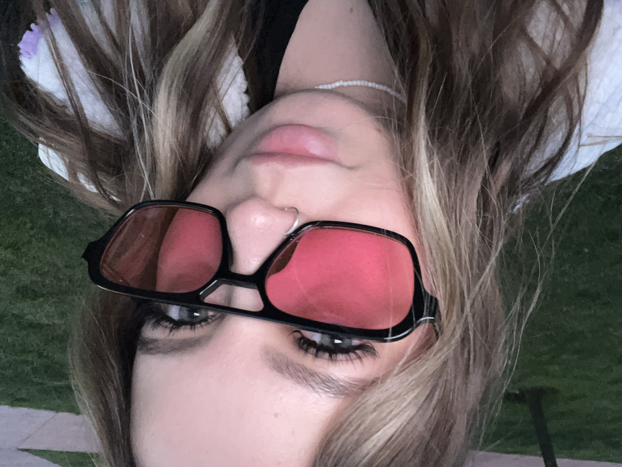 The image presents an upside-down, close-up portrait of a person, likely female, lying outdoors in Krasnodar, Russia. The subject has long, wavy brown hair with blonde highlights, which is spread out around their head. They are wearing distinctive red-tinted sunglasses with thick black frames, through which their green or blue-grey eyes and long eyelashes are visible. The person's full, pink lips are slightly parted, and a small silver septum nose ring adorns their nose. A white beaded necklace or choker is visible around their neck.
The background shows patches of green grass on either side of the person's head, indicating an outdoor setting, possibly a lawn or park. What appears to be a paved or concrete surface is subtly visible below some of the hair on the right side. The lighting is somewhat dim, suggesting the photograph was taken during evening or twilight hours. The person is looking directly at the camera, posing for the shot with a neutral expression. No visible text is present in the image.