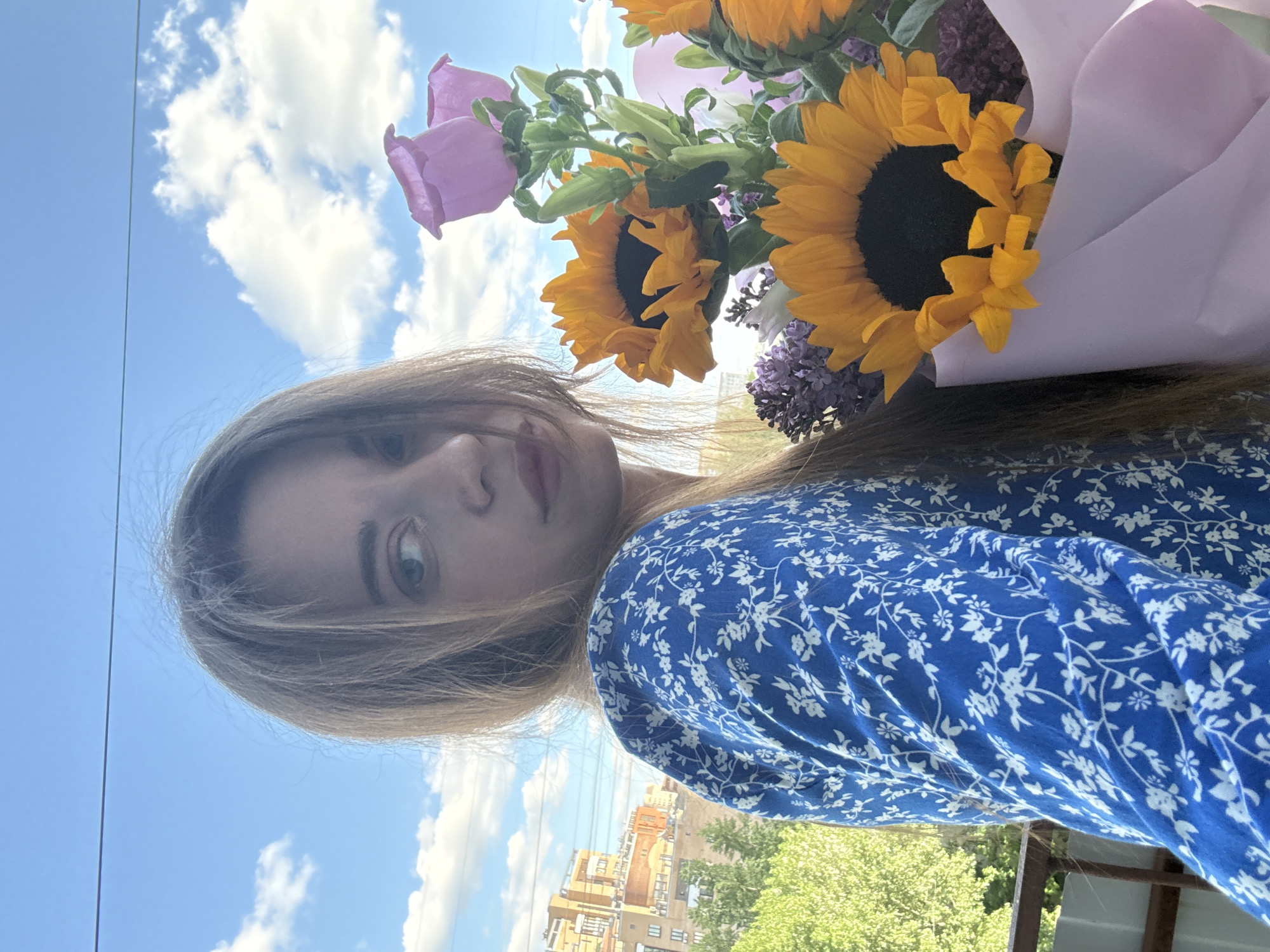 A young woman with long blonde hair is holding a bouquet of flowers. The bouquet contains sunflowers, purple bellflowers, and other flowers wrapped in light purple paper. The woman is wearing a blue dress with a small white floral pattern. In the background, there is a blue sky with scattered clouds. Buildings and trees can be seen in the distance. Based on the architectural style of the buildings, the location is consistent with Moscow, Russia.
