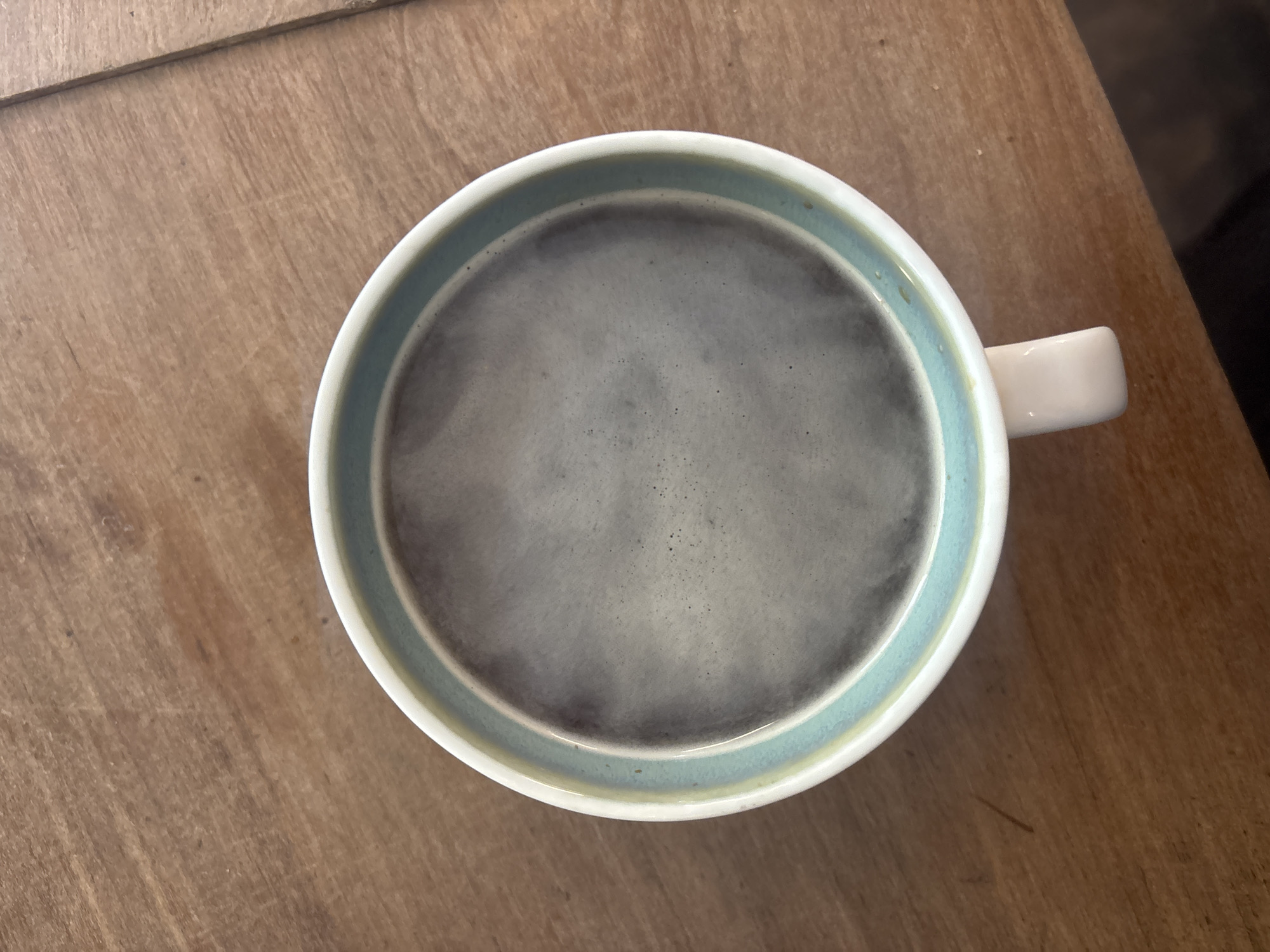 The image provides a top-down view of a single mug of coffee placed on a wooden surface. The coffee inside the mug is dark, and its surface displays a light gray, slightly foamy texture with numerous small bubbles and a subtle haze, suggesting it is hot.
The mug itself is primarily white or cream-colored on its exterior. Its inner rim features a decorative band: a prominent light blue-green stripe, underneath which a thinner, more subtle yellowish-green line is visible just above the coffee. A small, cream-colored handle protrudes from the right side of the mug. Small droplets or slight discolorations are visible on the outer edge of the mug's rim.
The wooden surface, likely a table or countertop, is a light to medium-brown tone. It exhibits clear wood grain patterns and horizontal lines, possibly indicating joins between planks or general wear. The lighting is even, suggesting an indoor setting during the day. There is no visible text within the image. While the provided context is Buenos Aires, Argentina, no specific visual elements in the media file indicate this geographical location.