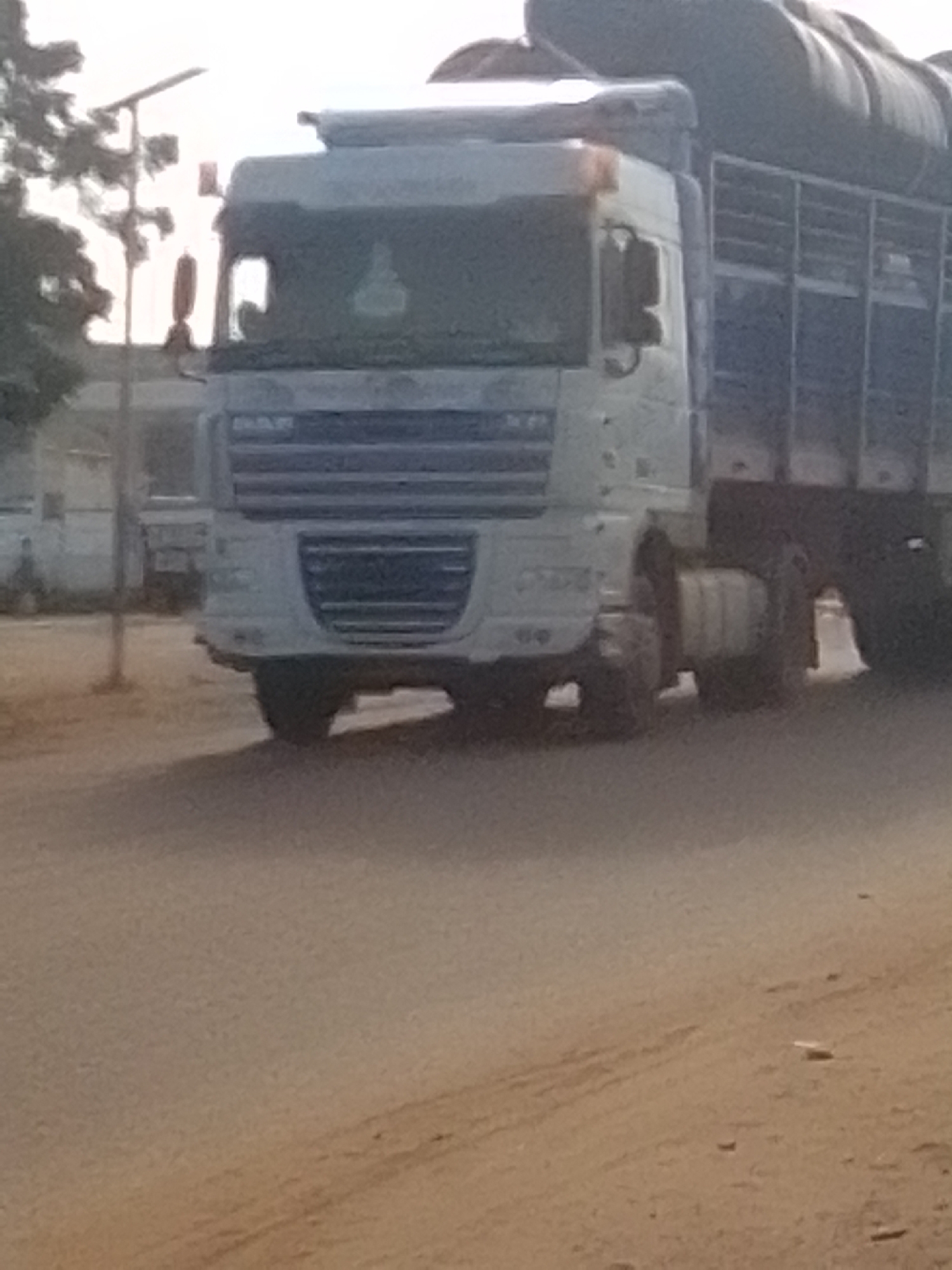 A white DAF truck with a large cargo trailer is parked on a dirt road in the city of Galadima, Nigeria. The truck is loaded with what appears to be hay or similar agricultural produce, piled high in the trailer. The truck's cab is white and shows some dirt and wear. The trailer is a dark color, possibly blue or black, and has visible vertical bars along its sides. The road surface is uneven and dusty, with some loose debris scattered around. In the background, there are trees and a few buildings, including what looks like a smaller white vehicle parked further down the road. A utility pole with a light fixture is visible to the left of the truck. The sky is overcast, suggesting a hazy or cloudy day. There are no people visible in the immediate vicinity of the truck.