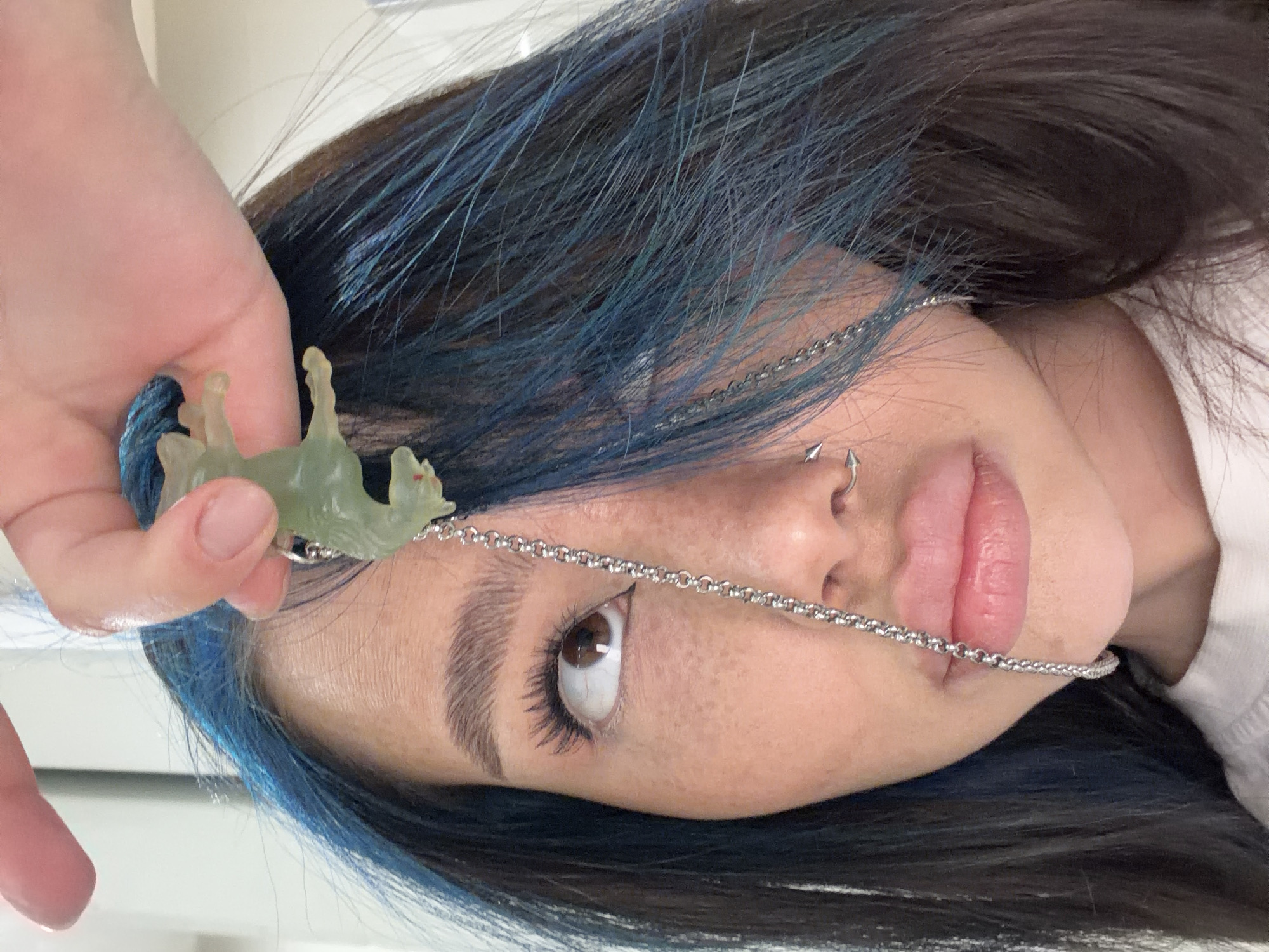 A close-up, rotated portrait features a young person, likely female, with dark hair strikingly highlighted with blue streaks. The individual has a light to medium skin tone, brown eyes, and prominent eyebrows. Freckles are visible across their nose and cheeks. Their lips are slightly parted, revealing a natural pink hue.
A delicate, silver-colored chain is draped across their face, extending from near the left eyebrow, over the bridge of the nose, and resting on the upper lip. A small, silver-colored piercing is visible in the left nostril, likely a septum piercing, and another small stud or piercing is on the side of the nose.
A hand with light skin and neatly trimmed nails holds up a translucent, light green, animal-shaped pendant—resembling a deer or similar quadruped with tiny red eyes—attached to a blue string or cord. The hand is positioned in the upper left of the frame. The subject's gaze is directed slightly upwards and to the right, conveying a calm and engaged expression.
The background is softly blurred, suggesting an indoor setting. A hint of a white garment, possibly a collar, is visible on the right side of the frame. The overall composition is a stylized portrait. No visible text is present in the image. The location context is Moscow, Russia.