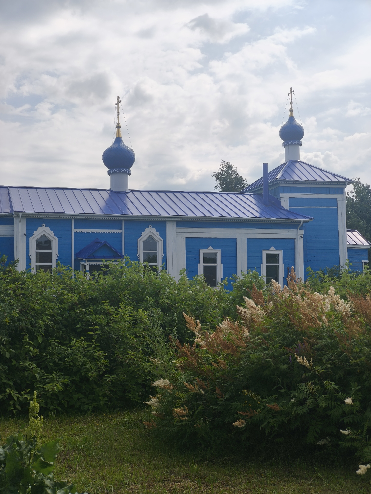 The image depicts the exterior of a blue wooden church with a blue metal roof and two blue domes, each topped with a gold cross. The church has white trim around its windows and along the roofline. In the foreground, there is lush green foliage and some flowering plants with white and beige plumes. The sky above is cloudy. The church is located in the village of Sokury, Russia.