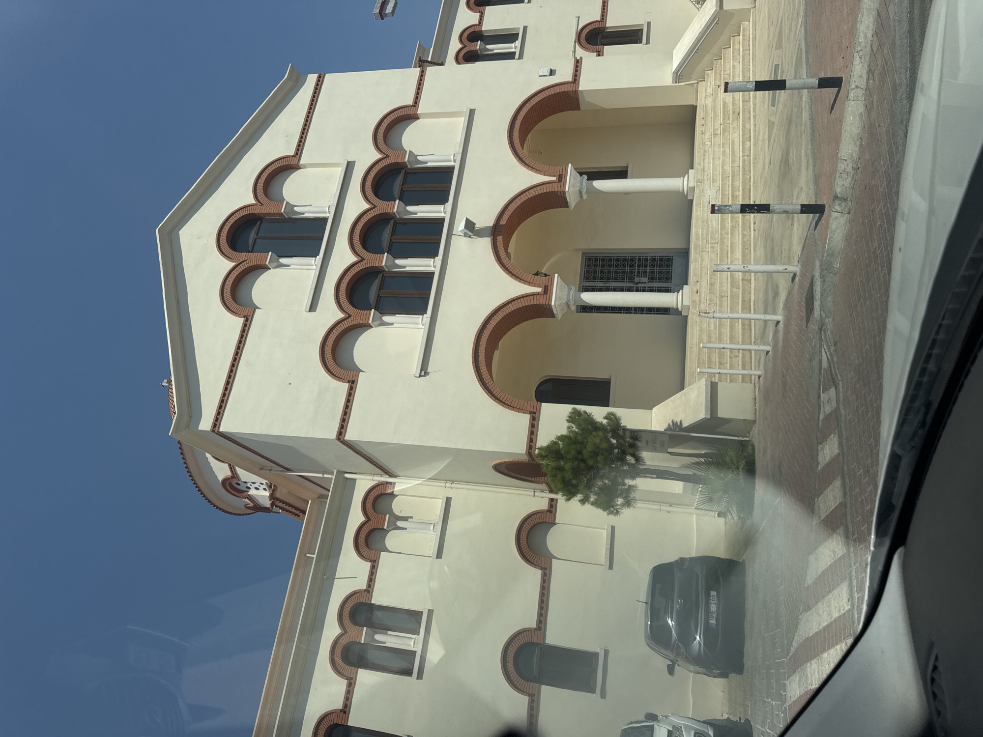 This is a daytime, exterior shot of a building with a light yellow facade and red brick trim, located in Limassol, Cyprus. The building features arched windows and doorways, with columns supporting a covered entrance area. A small evergreen tree stands to the left of the entrance. In the foreground, a dark gray car is parked. The ground is paved with red brick and white crosswalk markings. The sky is clear and blue. The scene appears to be a street view in front of a church or a similar public building.