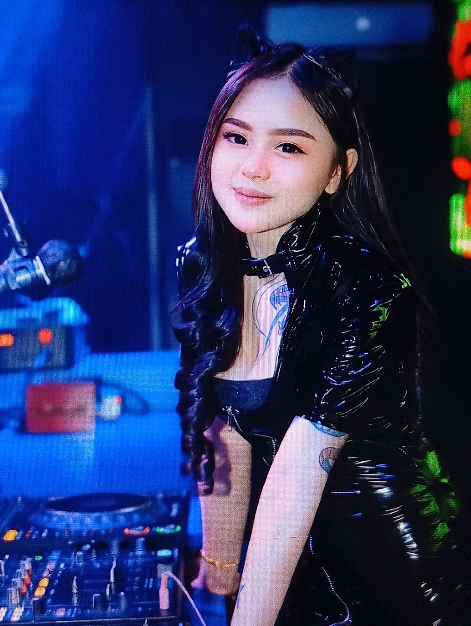 This is a close-up, medium-shot, horizontal, indoor photograph of a young woman with long, dark, wavy hair, wearing what appears to be a black, shiny, vinyl-like, form-fitting outfit. She has on black cat-ear-like accessories on her head. She has makeup on her eyes and lips. There is a tattoo visible on her upper chest area, and another on her right arm. She is wearing a gold bracelet on her left wrist.
She is positioned in front of a DJ setup, which includes turntables and mixers. The background is dark with areas of blue and green light, suggesting a club or performance venue. To the left of the woman, a microphone is visible, also part of the DJ setup. The overall lighting is dim and atmospheric, with the subject well-lit and in sharp focus.
There is no explicit information in the image to determine its specific location within the city of Lhokseumawe, Indonesia. However, the setting suggests a nightlife or entertainment establishment.