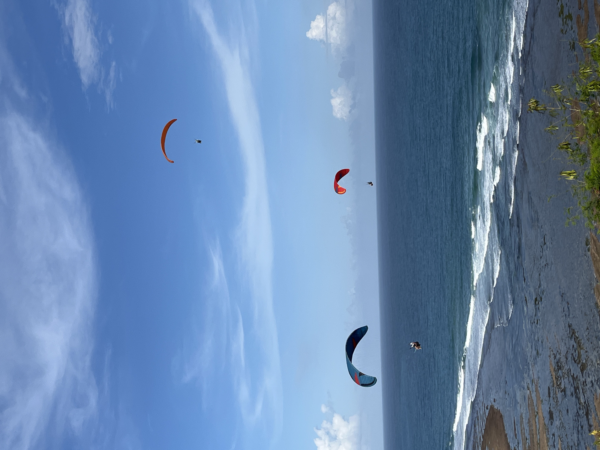 The image depicts a coastal scene, likely near a cliff overlooking the ocean in Kuta Selatan, Indonesia. Three paragliders are soaring in the sky with their pilots under their chutes. The sky is blue with scattered clouds. The ocean meets the land with breaking waves. The shoreline shows both sandy beach and exposed rocky reef. The upper-right corner shows some green vegetation atop the cliff.