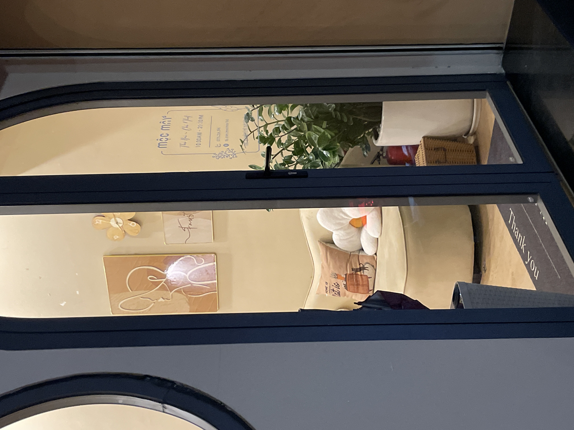 The image shows the interior of a shop in Da Lat, Vietnam, viewed through a glass door. The shop appears to be a cafe or a boutique. Inside, there is a beige sofa with several cushions. One cushion has an illustration of a cat and the text "HOME IS WHERE the CAT is". Another cushion is shaped like a white flower. On the wall, there are two framed pieces of art, one with abstract line art of faces and the other with script. There is also a small, light-colored flower decoration on the wall. A tall green plant in a white pot is positioned near the door. A small wicker basket and a red fire extinguisher are visible behind the plant. The floor has a mat with the text "Thank You" written on it. The shop has a sign with Vietnamese text that reads "mộc mây", followed by "The Hair - Chat Chat" and operating hours "10:00AM - 20:30PM". There is also a phone number and a Facebook page listed. The overall lighting suggests it is evening.