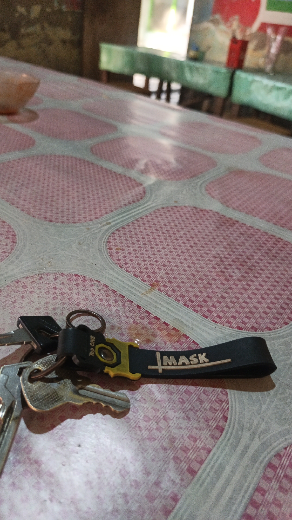 A close-up shot shows a set of keys on a pink and white patterned table. The keys are attached to a black lanyard with the word "MASK" printed in white. The lanyard also features a yellow accent piece. In the background, out of focus, are green tables with what appear to be chairs or stools underneath. The scene is indoors, possibly in a public eating area or food stall, given the table setting. The location is identified as Dawe, Indonesia, suggesting this is a scene from that region.