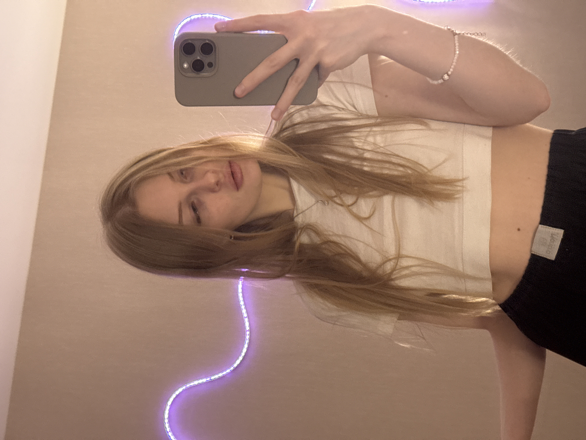 A mirror selfie captures a young female with long, straight blonde or light brown hair. She is lying down or angled such that her head is on the left side of the frame, facing slightly towards the camera with a neutral expression, her lips slightly parted. She is wearing a white cropped t-shirt and black bottoms, possibly shorts or sweatpants.
Her right arm is bent, holding a gray smartphone with a visible triple-lens camera system, taking the photo. A delicate, light-colored beaded bracelet with a small, sparkling charm is visible on her right wrist. In the background, a glowing purple LED strip light snakes across a plain, light beige or off-white wall. A small, light-colored fabric tag is visible on the black bottoms, with faint, partially obscured text that appears to read "LOUNGE" or "LOUNGEWEAR". The scene suggests an indoor setting, likely a bedroom or living space, in Perm, Russia.