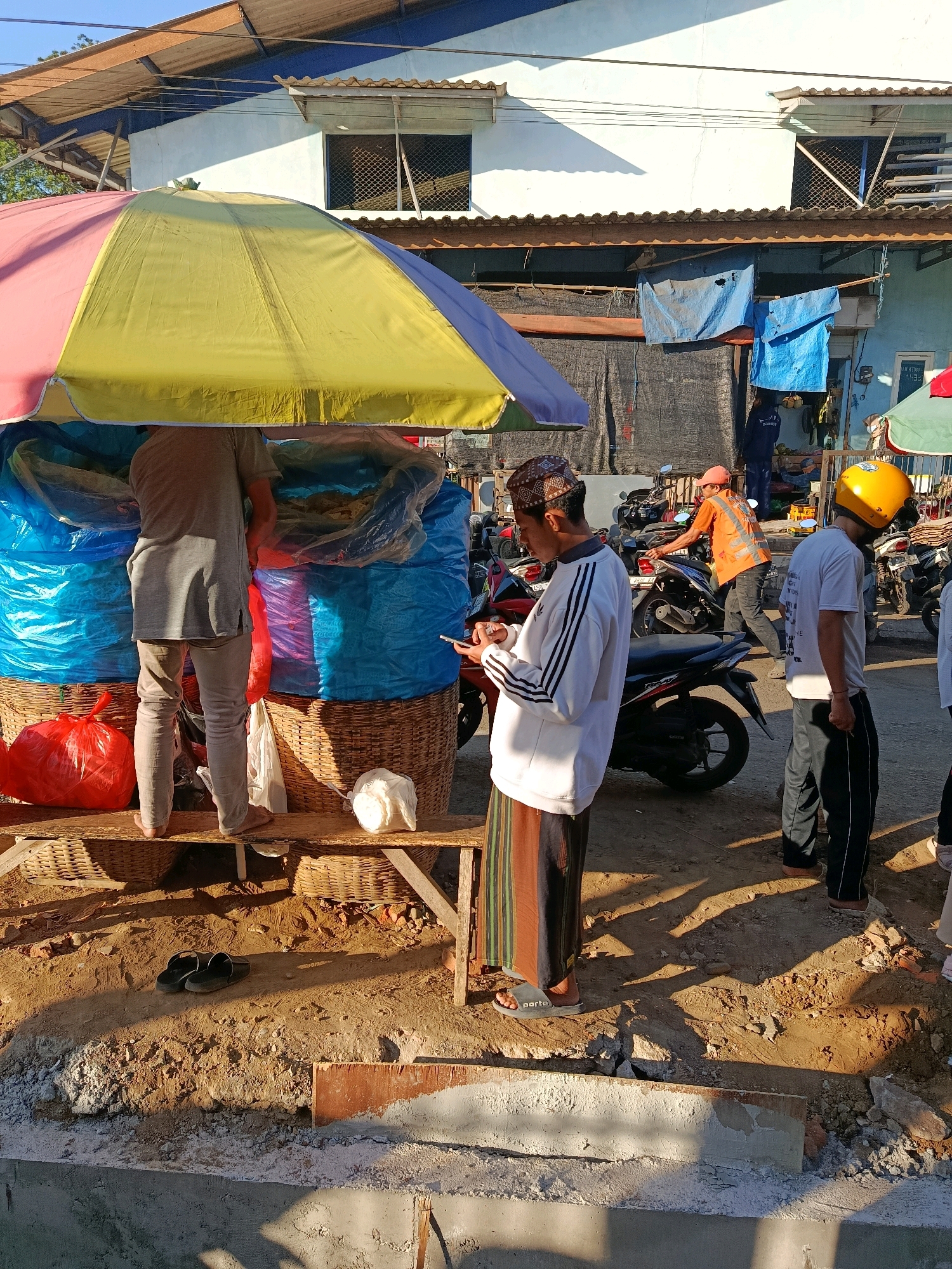 In Wandanpuro, Indonesia, under a colorful striped umbrella, a vendor stands barefoot behind a stall filled with large blue plastic-wrapped items and woven baskets. A red plastic bag is at the base of the stall, next to a white plastic bag and a white object. To the right, a young man in a white long-sleeved shirt and a patterned cap looks at his phone. Another man wearing a yellow helmet and a white t-shirt stands to his right. Behind them, motorcycles are parked and a person in an orange vest is interacting with one. The scene appears to be an outdoor market on a dirt surface, with buildings and tarps visible in the background.