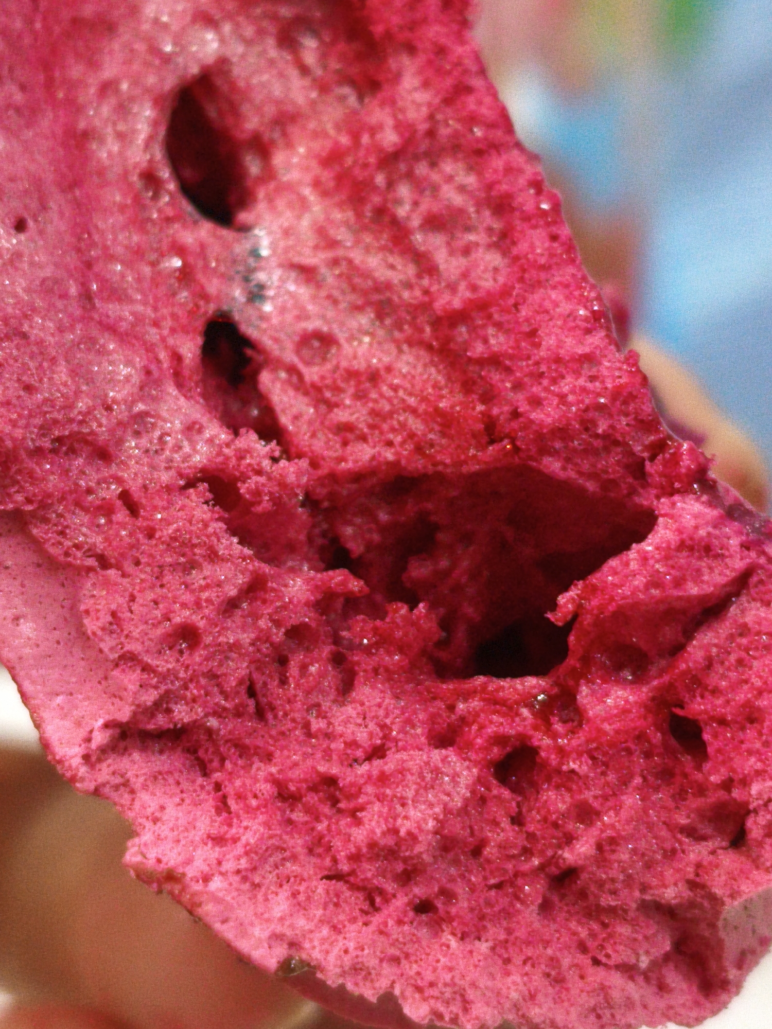 The image is a close-up, macro photograph of a brightly colored pink, porous substance. The texture appears somewhat frothy and irregular, with visible air pockets and a slightly shiny surface, suggesting it might be some kind of food or confection. The lighting is direct, highlighting the textured surface and creating small reflections. The background is out of focus and appears to be a gradient of light blue and some muted colors, possibly indicating an indoor environment with artificial lighting or a blurred outdoor scene. There is no discernible context of a specific location in Orenburg, Russia, or any identifiable people or actions. The focus is entirely on the detailed texture and vibrant color of the pink material.