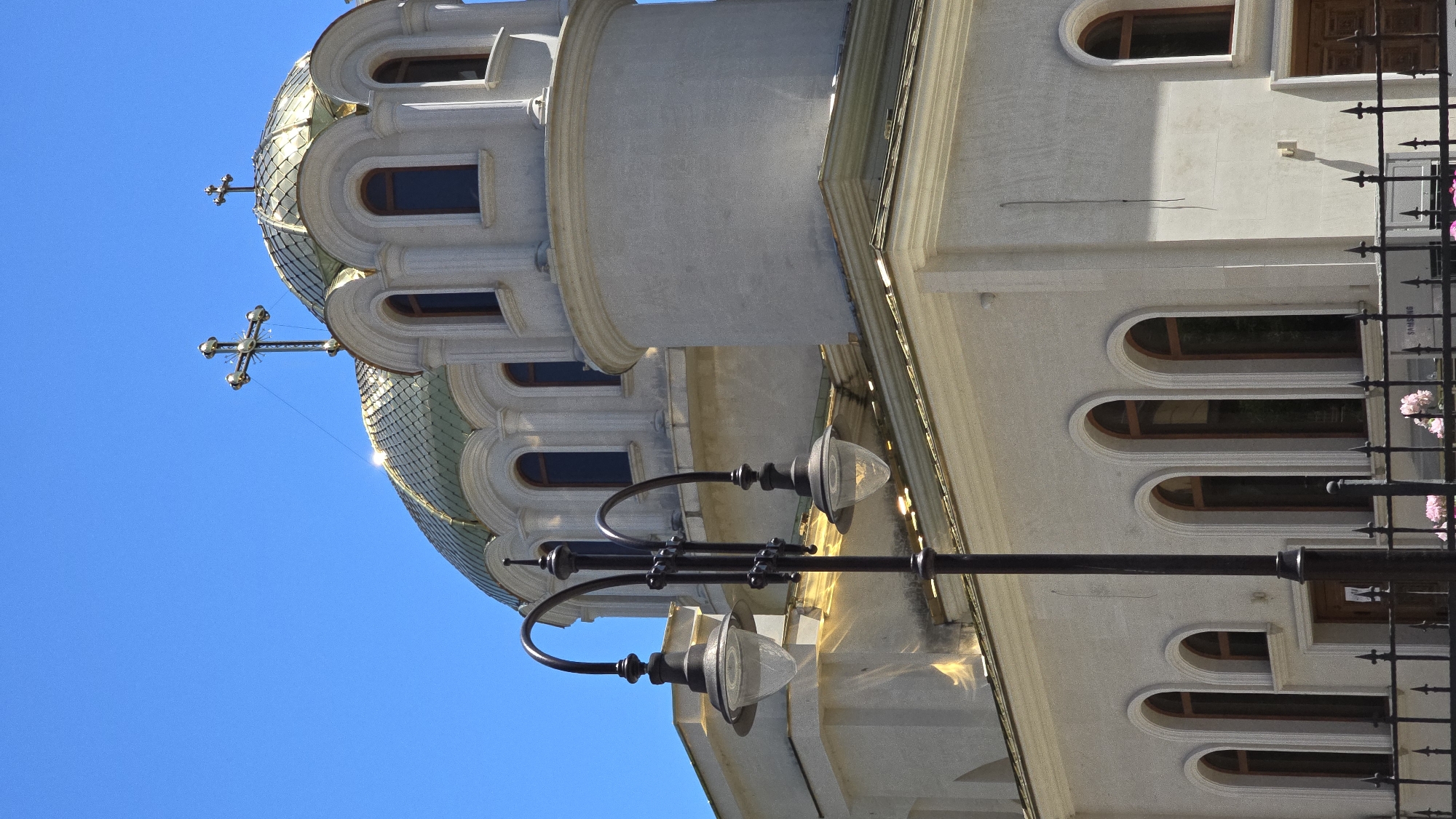 The image shows a partial view of the St. Nicholas the Miracleaker Church in Varna, Bulgaria. The church is a prominent landmark, characterized by its imposing architecture, including golden domes and a tall bell tower. The scene captures the church under a clear blue sky, with sunlight reflecting off its gilded surfaces. In the foreground, a black lamppost with two lamps stands, partially obscuring the view of the church's facade. The church features arched windows and decorative stonework. A black metal fence runs along the bottom of the image, with some pink flowers visible behind it. The overall impression is one of grandeur and religious significance, set against a bright, sunny day.