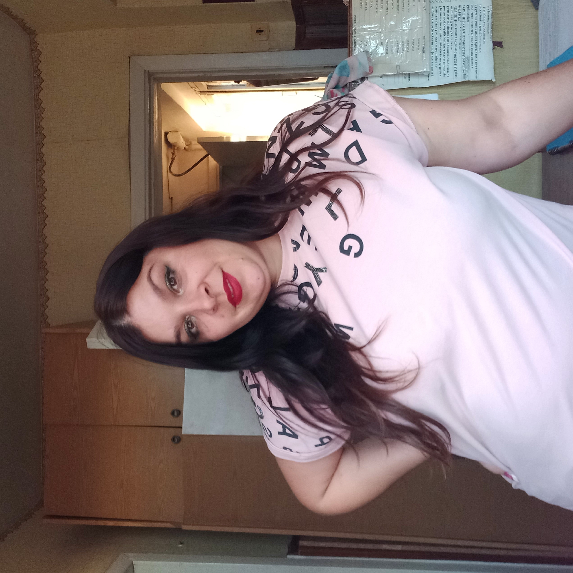 A medium shot shows a woman standing indoors, her torso angled toward the camera. She has long, dark brown hair that falls past her shoulders and wears makeup, including bright red lipstick and eyeliner. Her short-sleeved, pale pink t-shirt is adorned with black letters and shapes. Her left arm is bent at the elbow and rests on her hip, while her right arm is partially visible in the lower right corner of the frame. The background features wooden cabinets on the left and a doorway leading to another room on the right. The walls are covered in a light-colored wallpaper with a subtle pattern, and a decorative border runs along the top. The lighting suggests an indoor environment. The overall scene appears to be within a residential setting, potentially a kitchen or living area, in an unspecified location in Ukraine.