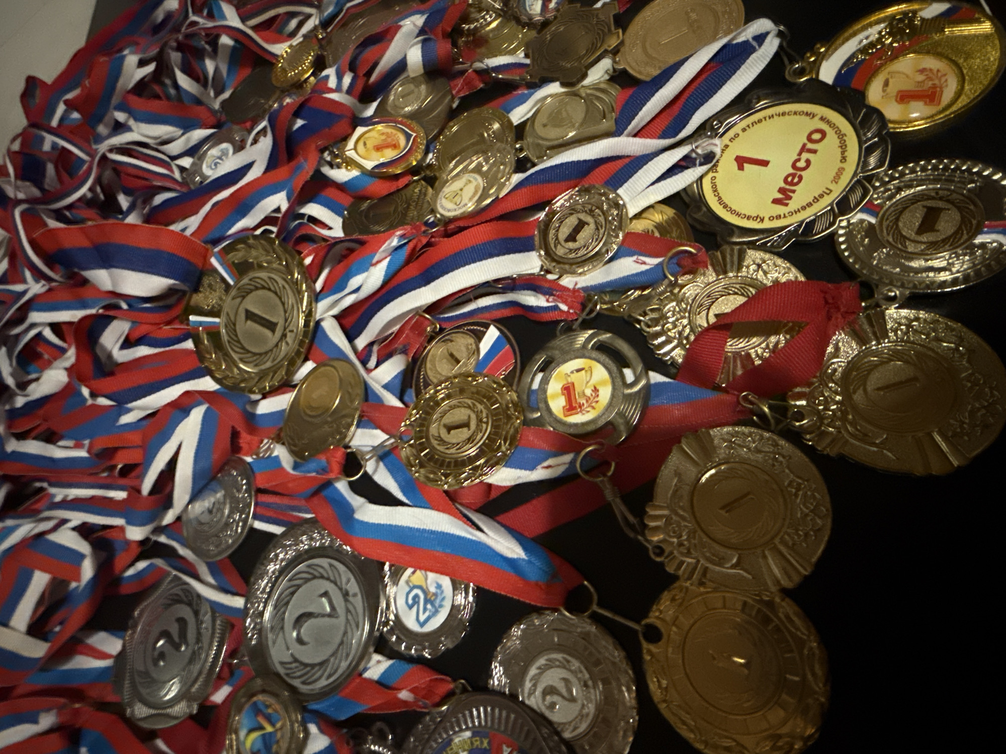 The image displays a pile of sports medals with ribbons in the colors of the Russian flag (red, white, and blue). The medals are of varying sizes and metallic finishes, primarily gold and silver, and are inscribed with numbers (1, 2) denoting placement. Some medals are of newer manufacture, while others are older. One particular medal includes the words "1 место" (first place) in Russian, and "Первенство Красносельского района по атлетическому многоборью 2009 года" (Krasnoselsky district championship in track and field decathlon 2009 year). The location could be Krasnoselsky district in St. Petersburg, Russia, or a place where sports medals from St. Petersburg are stored. The medals appear to be randomly strewn together on a dark surface.