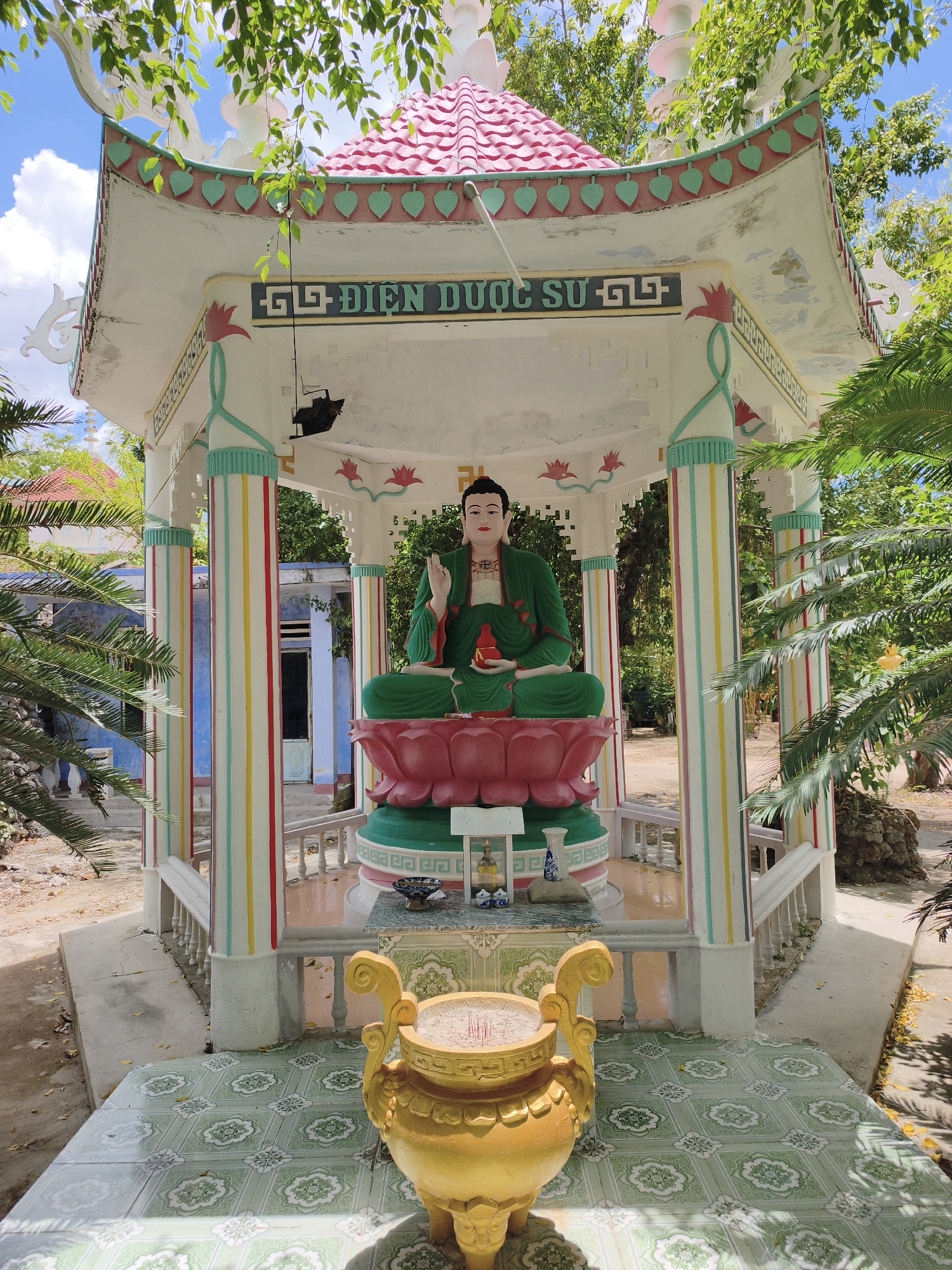 The image shows a colorful, outdoor shrine dedicated to Buddha. The central subject is a large, seated Buddha statue adorned in green robes, resting on a pink and green lotus flower base. The statue is housed within a white, open-sided structure with a pink, tiled, multi-tiered roof decorated with green leaf motifs and ornate white trim. The structure is supported by pillars painted with vertical stripes of red, yellow, green, and blue. In front of the shrine, on a tiled floor, sits a large, ornate, golden incense burner filled with incense sticks. The shrine is surrounded by lush green foliage, including palm trees, and a bright blue sky with scattered clouds is visible above. In the background, a simple blue building with a white door can be seen, suggesting a rural or semi-rural setting. The overall impression is one of a peaceful and culturally significant religious site. Based on the architectural style and the script on the banner above the Buddha statue, this shrine is likely located in Cam Ranh, Vietnam.