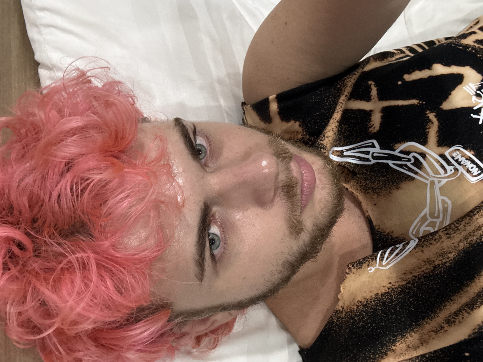 A man with pink hair and a beard lies on a white bed. He is wearing a black and tan tie-dye shirt with white graphic designs. He has blue eyes and fair skin. The scene appears to be indoors. The location in Đà Nẵng, Vietnam is not determinable from the image.