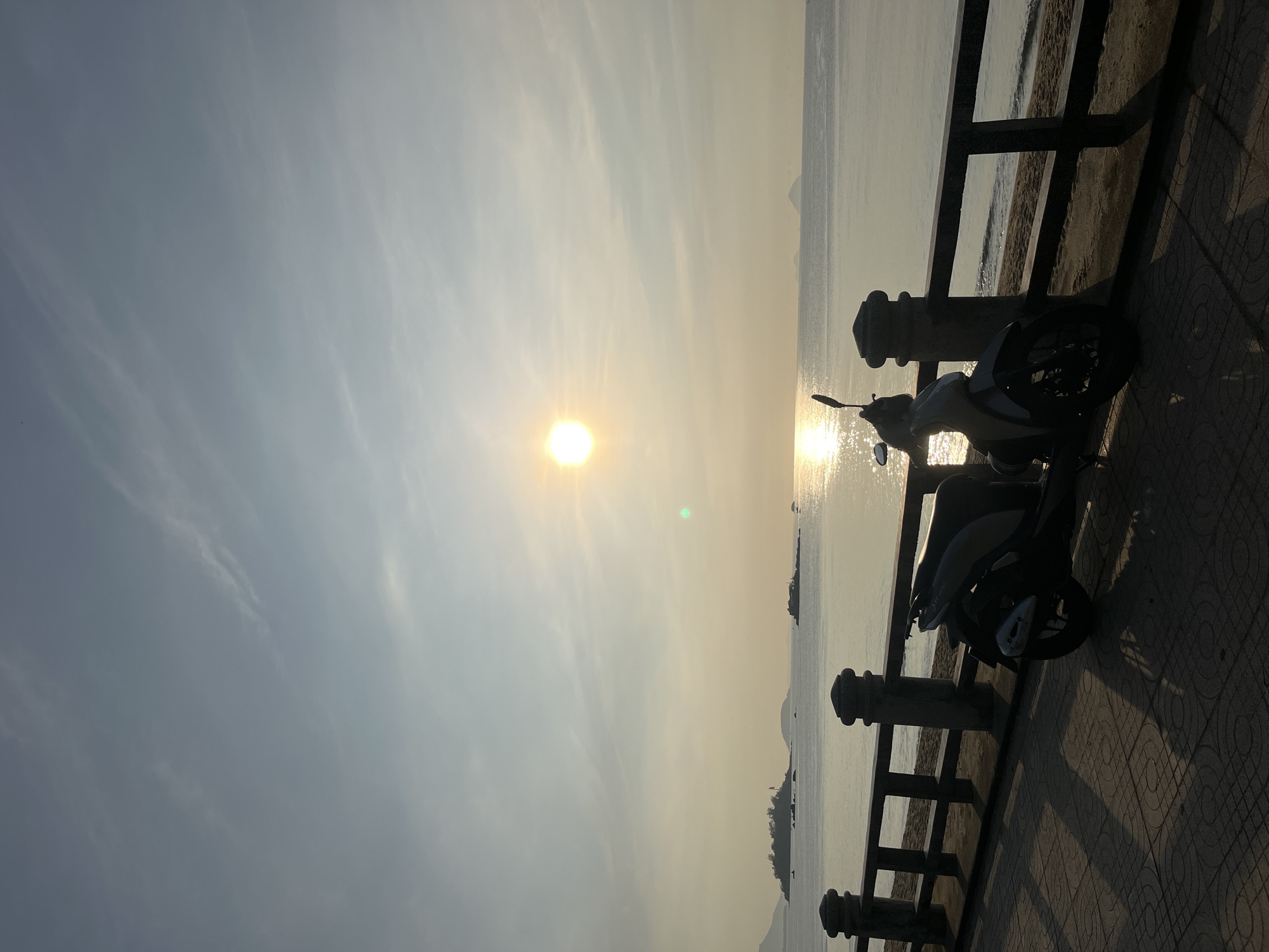 The image shows a motorcycle parked on a tiled walkway alongside a black fence near a beach. The sun is setting over the water, creating a bright reflection on the sea. The location appears to be in Nha Trang Ward, Vietnam, as evidenced by the coastal environment and the type of motorcycle commonly found in the area. There are distant islands visible on the horizon. The scene suggests a calm, possibly touristic environment, with the motorcycle likely belonging to a local or tourist.