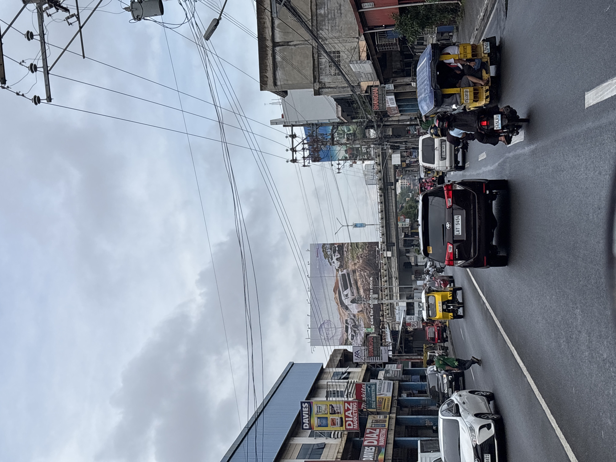 The image shows a street scene in Davao City, Philippines, on a cloudy day. The road is wet, indicating recent rain. Several vehicles are present, including a white Toyota Vios, a black Toyota Innova, a yellow tricycle, and a motorcycle. A man in a green shirt is crossing the street. Buildings line the street, featuring various signs and advertisements, including a large billboard for Nissan. Power lines are visible overhead, contributing to the urban landscape. The overall impression is of a busy, typical urban environment in the Philippines.