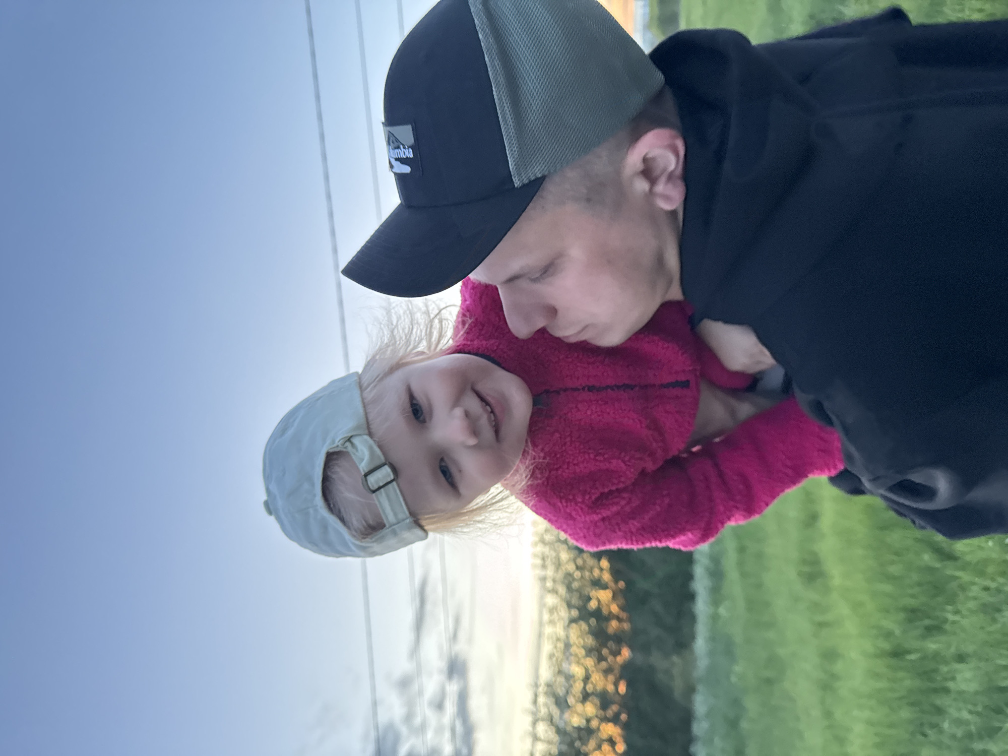 A man and a young child are depicted in a close-up shot. The man is wearing a black and olive green baseball cap with a small logo on the front. He has short, light-colored hair. He is wearing a black hoodie. The child is wearing a light gray baseball cap and a bright pink fuzzy jacket. The background is a mix of sky, blurred vegetation, and what appears to be an outdoor setting. The precise location in Горный, Russia, cannot be determined from the image.