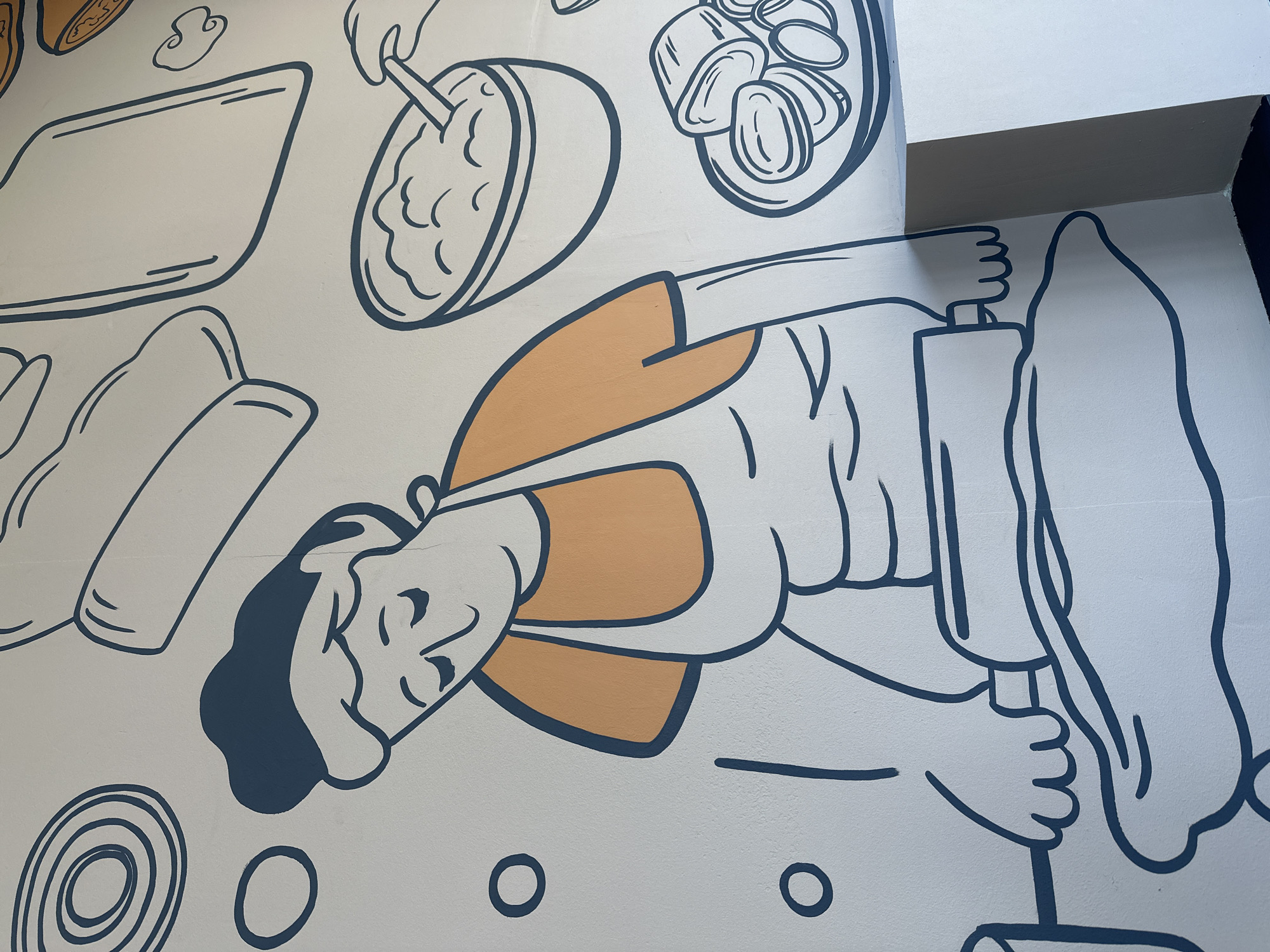 A mural depicts a stylized cartoon cook rolling out dough. The cook, rendered in navy blue and tan, is depicted mid-action, wielding a rolling pin. Above the cook are various outlined images of food and cooking tools. The background is white. The location, Votkinsk, Russia, is not definitively evident from the image itself, but can be inferred if given the location of the mural.