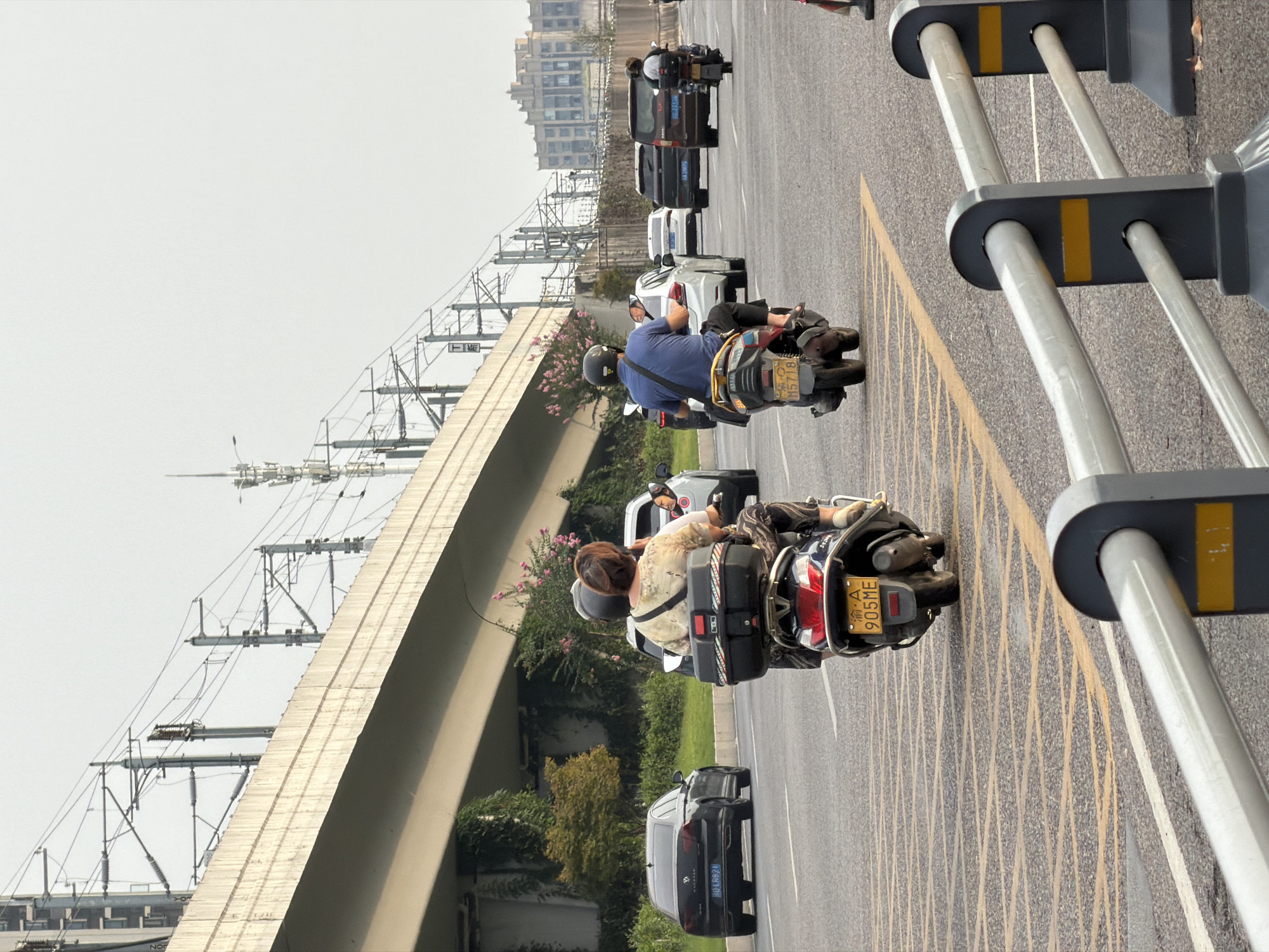The image captures a street scene in Jiangjin, China, featuring a multi-lane road with various vehicles and infrastructure. In the foreground, a metal guardrail with yellow markers is visible, partially obscuring the road. The road itself has yellow markings, indicating lanes and potential hazard areas.
Several vehicles are present on the road. Two individuals are riding on a scooter in the left lane, with a passenger seated behind the driver. The scooter has a black storage box mounted on the rear and a yellow license plate. Further ahead in the same lane, a black sedan is driving. In the right lane, another scooter is traveling, carrying a rider wearing a blue shirt and a helmet, also with a storage box and a yellow license plate. Ahead of this scooter, other vehicles including a white sedan, a dark SUV, and a brown minivan are discernible.
Overhead, a large concrete overpass stretches across the frame, supported by pillars. Electrical wires and poles, likely for a railway system, are suspended above the overpass. Green foliage and flowering plants are visible on the embankment beneath the overpass. In the background, a building with a distinctive architectural style is situated, along with what appears to be a cell tower. The sky is overcast, suggesting diffused daylight.
