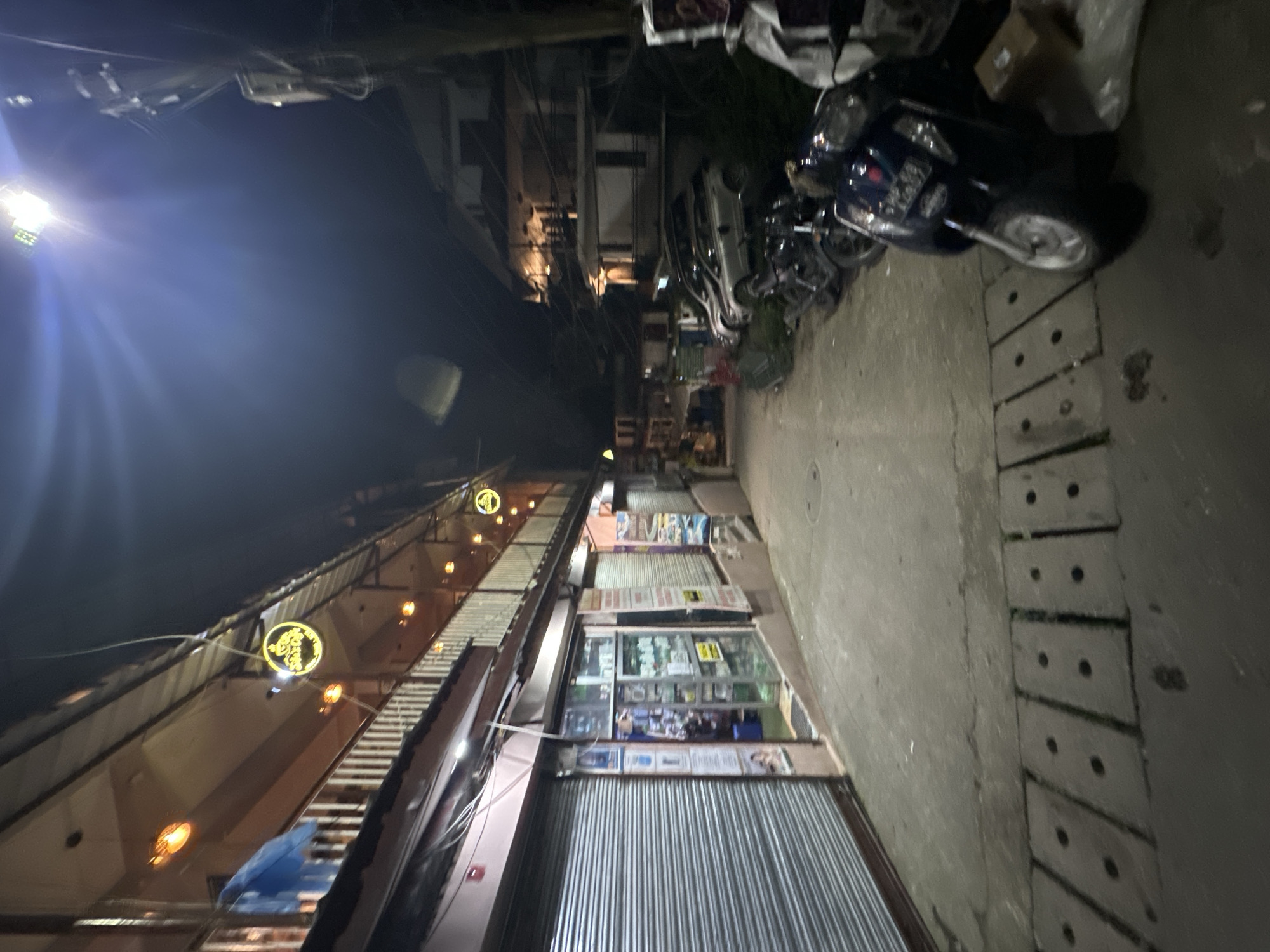 The image shows a narrow street at night, possibly in a market or residential area, with buildings on both sides. The street is paved and has some visible drains. On the left side, there are shops with their shutters down, and an upper floor with balconies and glowing lanterns. One circular neon sign with text is visible on the building. On the right side, there are more buildings, some appearing to be multi-story, and several parked motorcycles and cars. A bright streetlight illuminates the scene from above and to the left. The overall atmosphere is dimly lit, suggesting it's late at night or early morning.