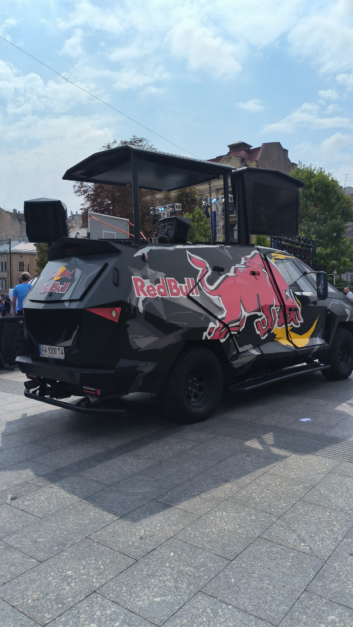 A black, heavily armored vehicle with the Red Bull logo and camouflage pattern is parked on a city square in Lviv, Ukraine. The vehicle is equipped with large speakers mounted on top and on the sides, suggesting it's used for events or promotions. The license plate reads "AA 9200 TA". In the background, buildings with distinctive architecture and trees are visible under a cloudy sky. A few people are present, including one in a blue shirt standing to the left of the vehicle, and another partially visible on the right. The ground is paved with large, grey rectangular tiles.