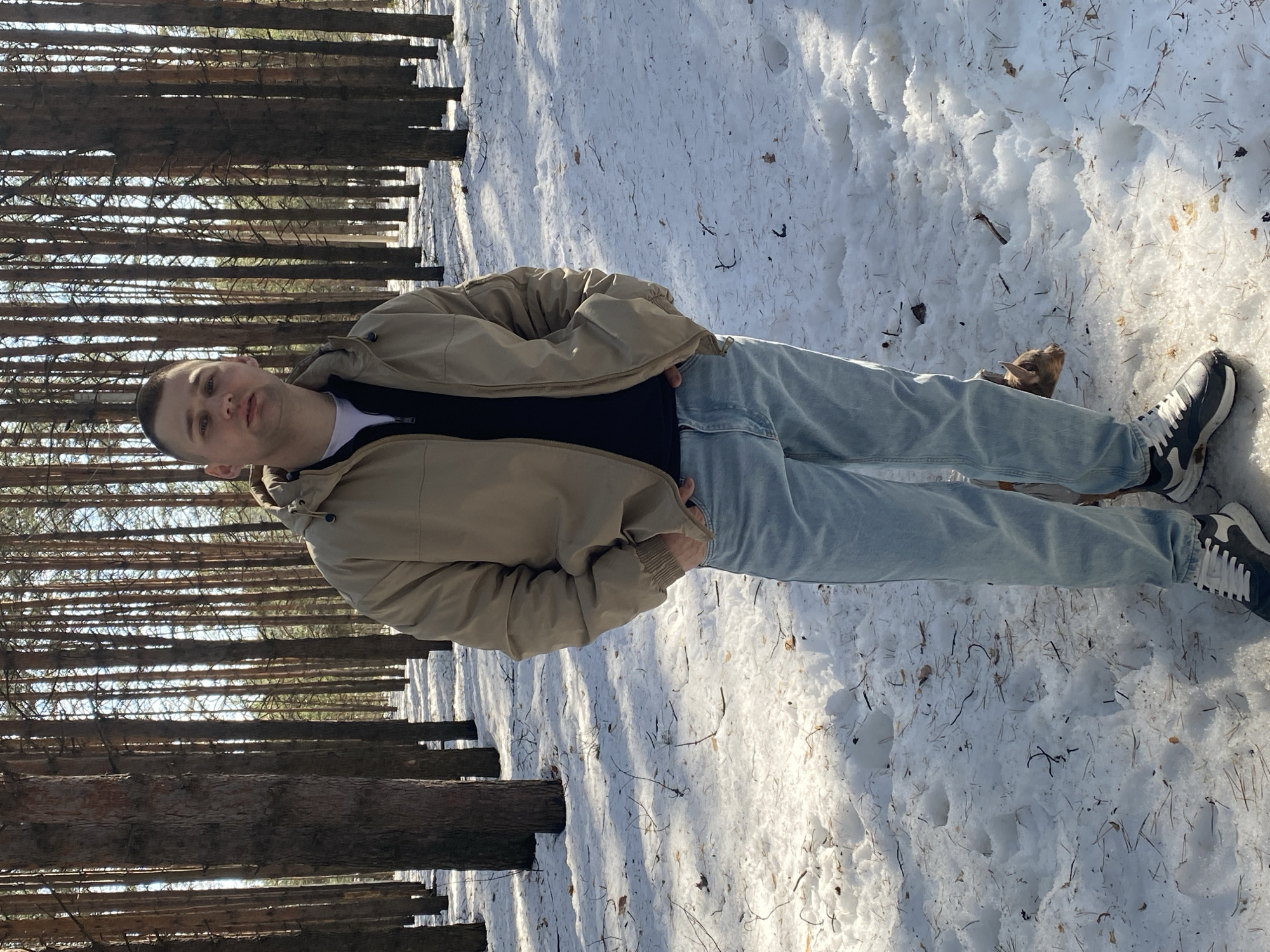 A male individual stands in a snow-covered pine forest located in Chaikovsky, Russia. The man, appearing to be in his twenties or thirties, has a short haircut and a neutral expression while looking directly forward. He is wearing a tan hooded jacket over a dark, possibly navy or black, quarter-zip shirt, light blue jeans, and dark athletic shoes with white soles and laces. Both hands are placed in the pockets of his jacket. A small, light brown-furred dog is partially visible on the ground behind his left leg. The forest floor consists of patchy snow, interspersed with exposed earth, pine needles, and twigs. Numerous tall, slender pine trees with dark brown trunks extend into the background, indicating a dense woodland environment under natural daylight.