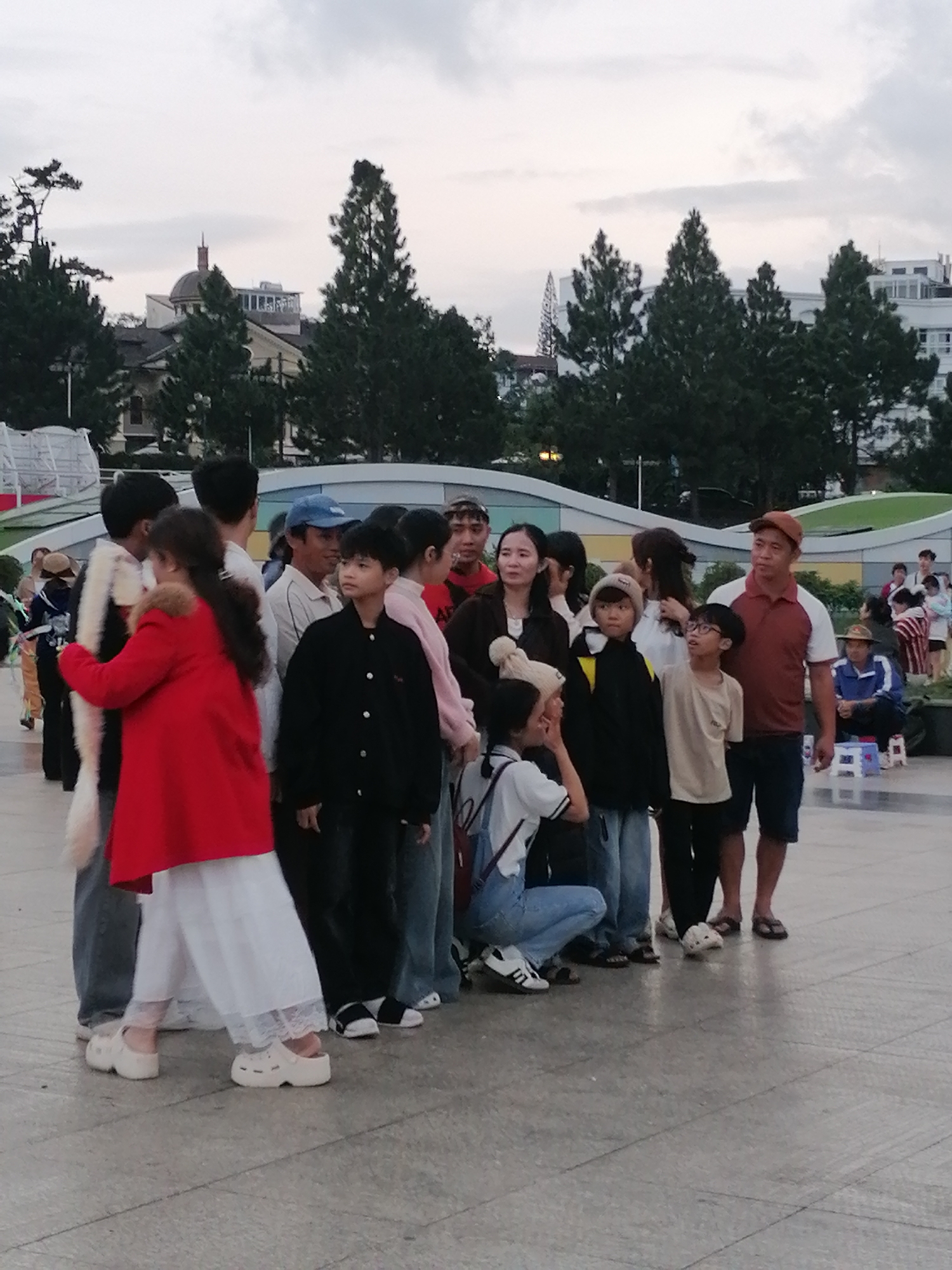 The image depicts a group of people, likely tourists, gathered in an outdoor public space in Da Lat, Vietnam. In the background, there are tall pine trees and buildings, including a domed structure and modern architecture, suggesting an urban or semi-urban setting. The foreground shows a paved ground, possibly a plaza or walkway, with a group of individuals of various ages, including children and adults, standing together. Some are looking in different directions, while others appear to be interacting. The lighting suggests it might be overcast or late afternoon. The people are dressed in casual clothing, and one individual is wearing a distinctive red coat. The overall impression is of a leisurely gathering or waiting in a tourist area.