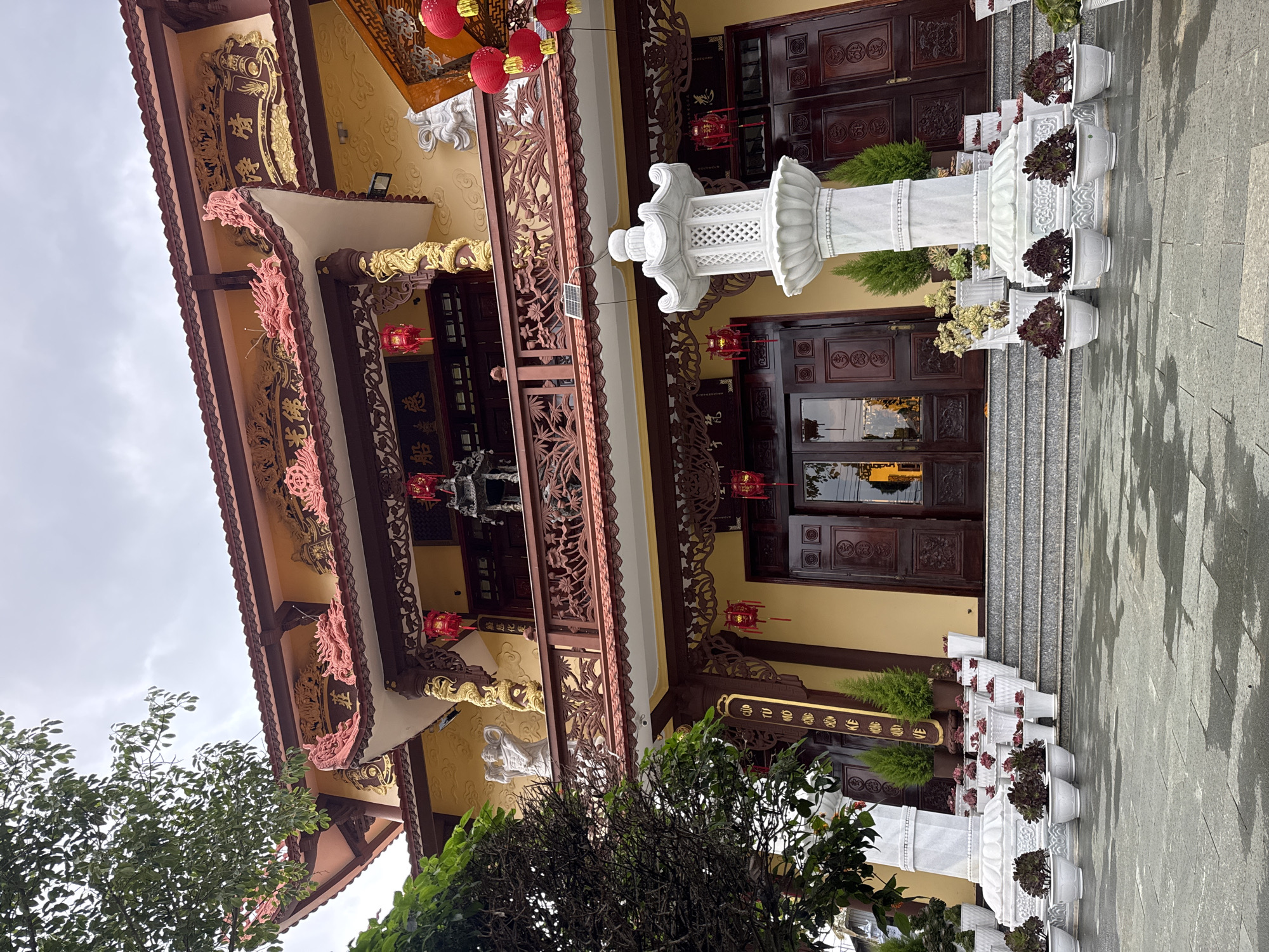 A low-angle shot depicts a multi-story temple-like building in Da Lat, Vietnam. The building features traditional East Asian architectural elements, with intricate wood carvings and decorations, red lanterns, and a yellow facade. Dark brown wooden doors with circular patterns are visible at the entrance. A tall, white stone pillar stands in front of the building, flanked by rows of potted plants on the steps leading up to the entrance. The foreground is partially obscured by a tree with green foliage. The sky is overcast, casting a soft light on the scene. The paving stones on the ground appear wet.