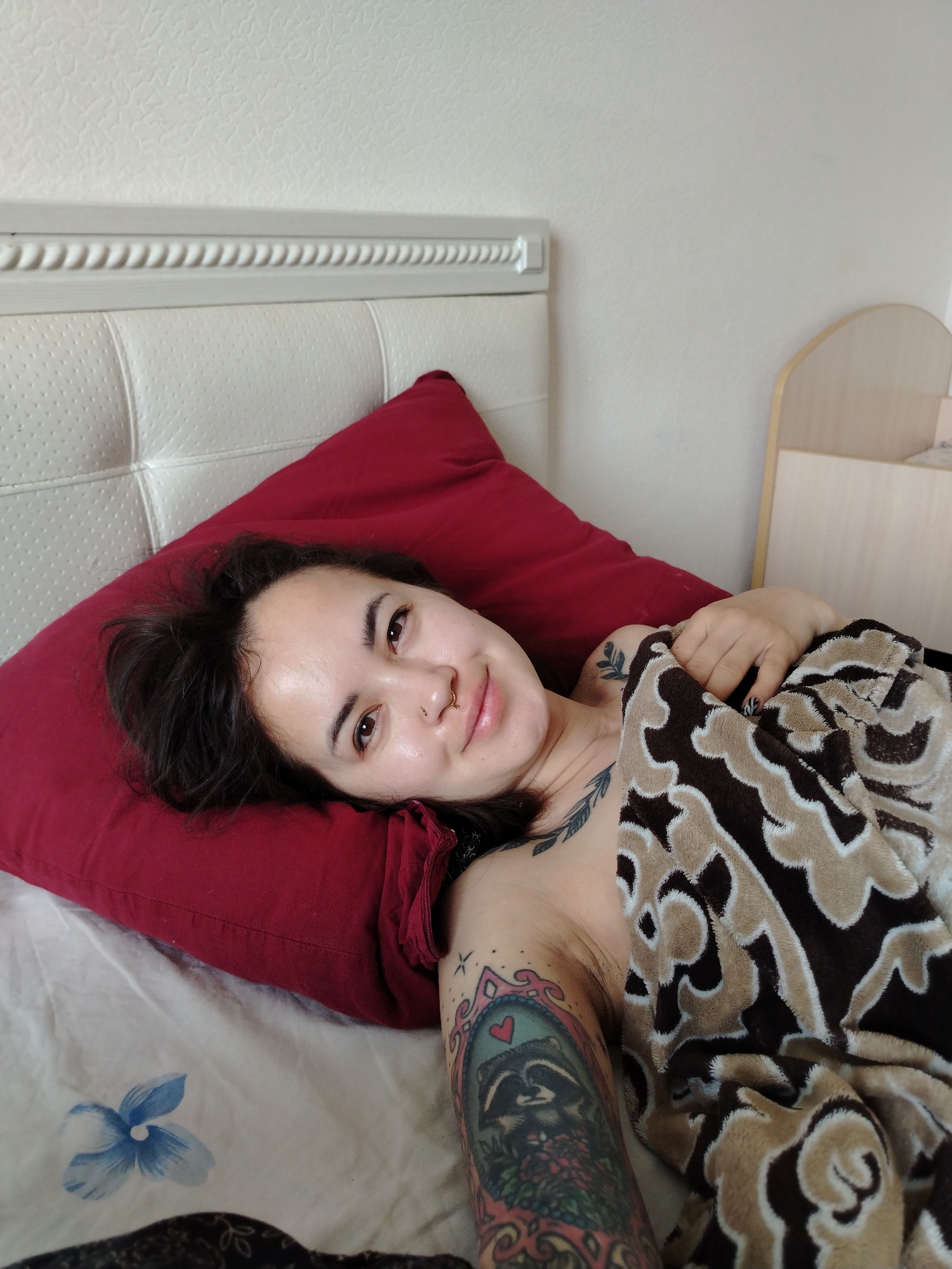 A person is lying in bed. The person has dark hair, fair skin, and a nose ring. They are lying on a red pillow against a white headboard. They are partially covered with a patterned blanket in shades of brown. The person has multiple tattoos on their arms and chest. The bed has white sheets with a blue floral pattern. The background is a plain white wall and a light wood-colored nightstand. The location is not depicted in the photo.