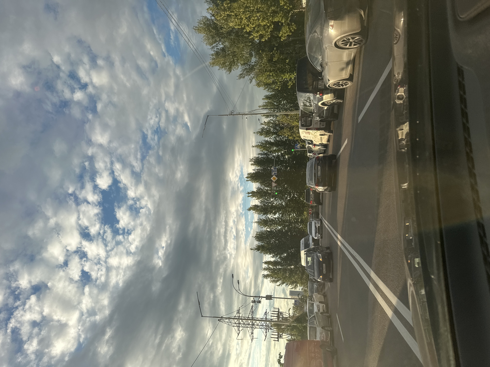 This is a photograph taken from the perspective of a driver in Kyiv, Ukraine. The scene depicts a busy highway with multiple lanes of traffic. Cars are bumper-to-bumper, indicating a traffic jam. The weather appears to be partly cloudy with dramatic cloud formations and patches of blue sky. On the left side of the road, there are tall power lines and pylons, a common sight in urban environments. Trees line the right side of the road, with lush green foliage. The road itself is asphalt, marked with white lines, and the sun is casting long shadows, suggesting it is either early morning or late afternoon. The overall impression is a typical urban commuting scene in Kyiv on a cloudy day.