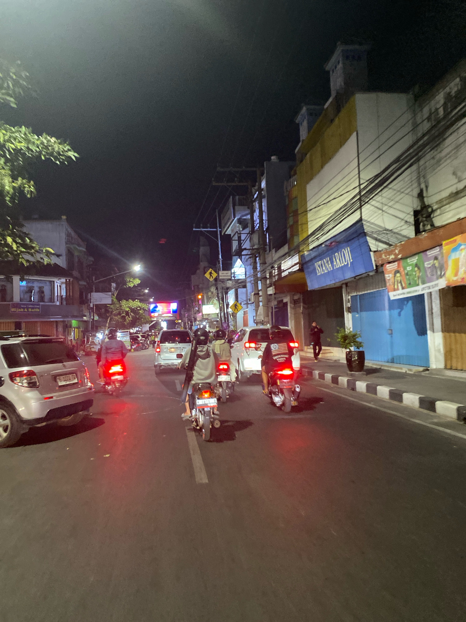 A nighttime, street-level, medium-shot captures a busy city street in Kediri City, Indonesia. The scene is illuminated by artificial light sources, including streetlights, building lights, and the headlights and taillights of vehicles. The road is paved and appears wet or reflective, with a white line dividing the lanes.
In the foreground, a silver SUV is parked on the left side of the road, facing away from the viewer. Its taillights are illuminated red. To the right of the SUV, a motorcycle with a rider is moving away from the viewer. The motorcycle's taillight is also red.
Further down the street, there is moderate vehicular traffic, primarily consisting of motorcycles. Several motorcycles, with riders, are visible moving in the same direction as the motorcycle in the foreground. Their taillights are illuminated red. Two white vans are also present in the traffic.
On the right side of the street, a sidewalk is visible, bordered by a curb with black and white markings. Buildings line the right side of the street. One building has a blue banner with white text that reads "ISTANA ARIOLI". Another building has a yellow facade. Power lines crisscross overhead.
On the left side of the street, more buildings are visible, some with illuminated windows. One building has a sign that reads "Hijab & Batik". Trees with green foliage are present on the left side of the street, partially obscuring the view of some buildings. Pedestrian crossing signs are visible on poles along the right side of the street.
A few individuals are visible, mostly on motorcycles, wearing helmets. One person, dressed in black, is walking on the sidewalk to the right of the street. The overall atmosphere is one of urban nighttime activity and traffic.