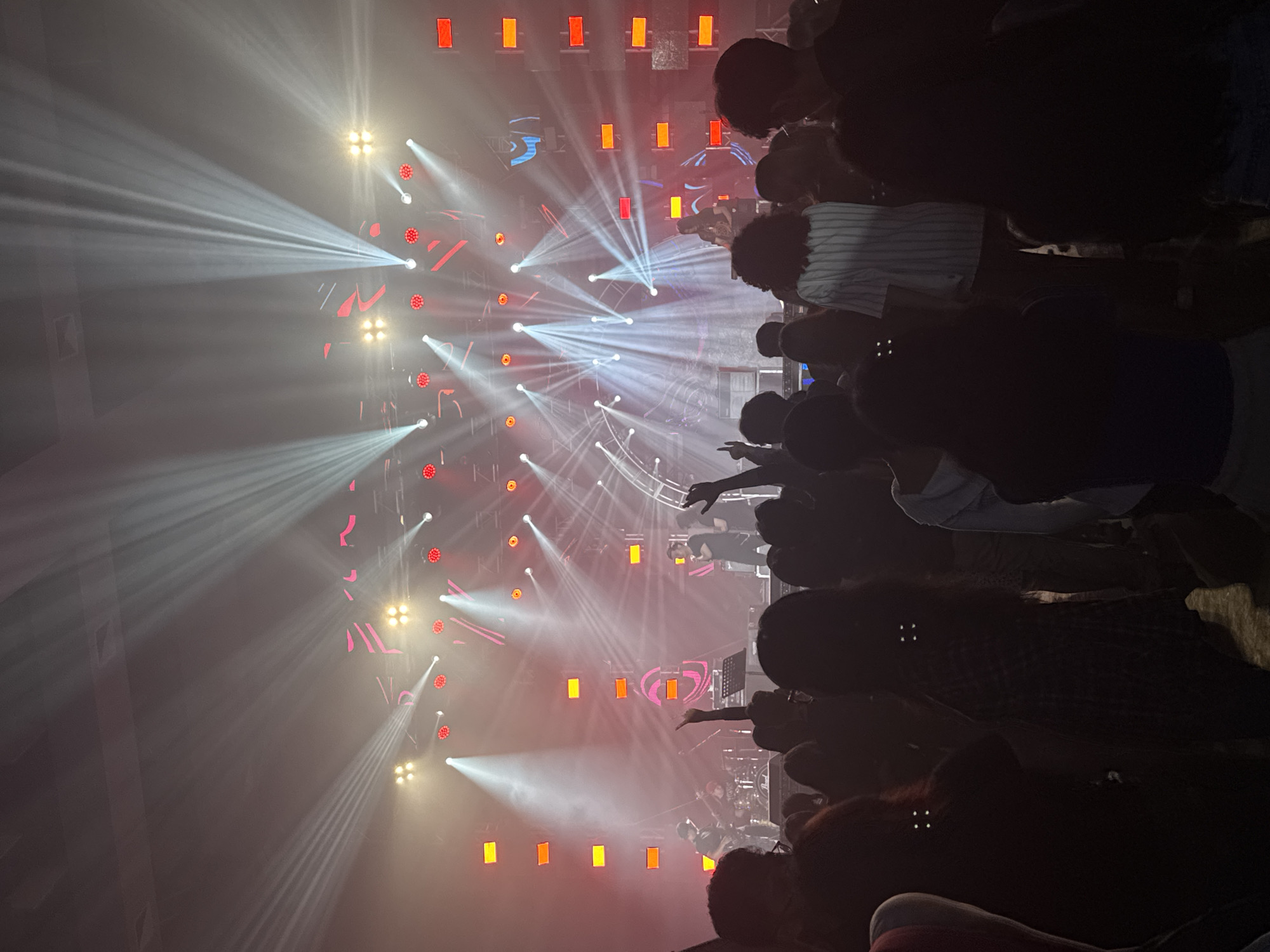 The image shows a live music performance at a concert venue, likely indoors, though the exact location in Davao City, Philippines cannot be determined from the image. A stage is set up with an elaborate lighting system featuring multiple spotlights and LED panels. White and colored lights shine across the venue, creating a dynamic visual display. A band is performing on stage, although the performers are mostly obscured by the stage lights and crowd. The audience is visible in the foreground, with many people raising their arms in the air, appearing to be enjoying the show. The scene suggests an energetic concert atmosphere.