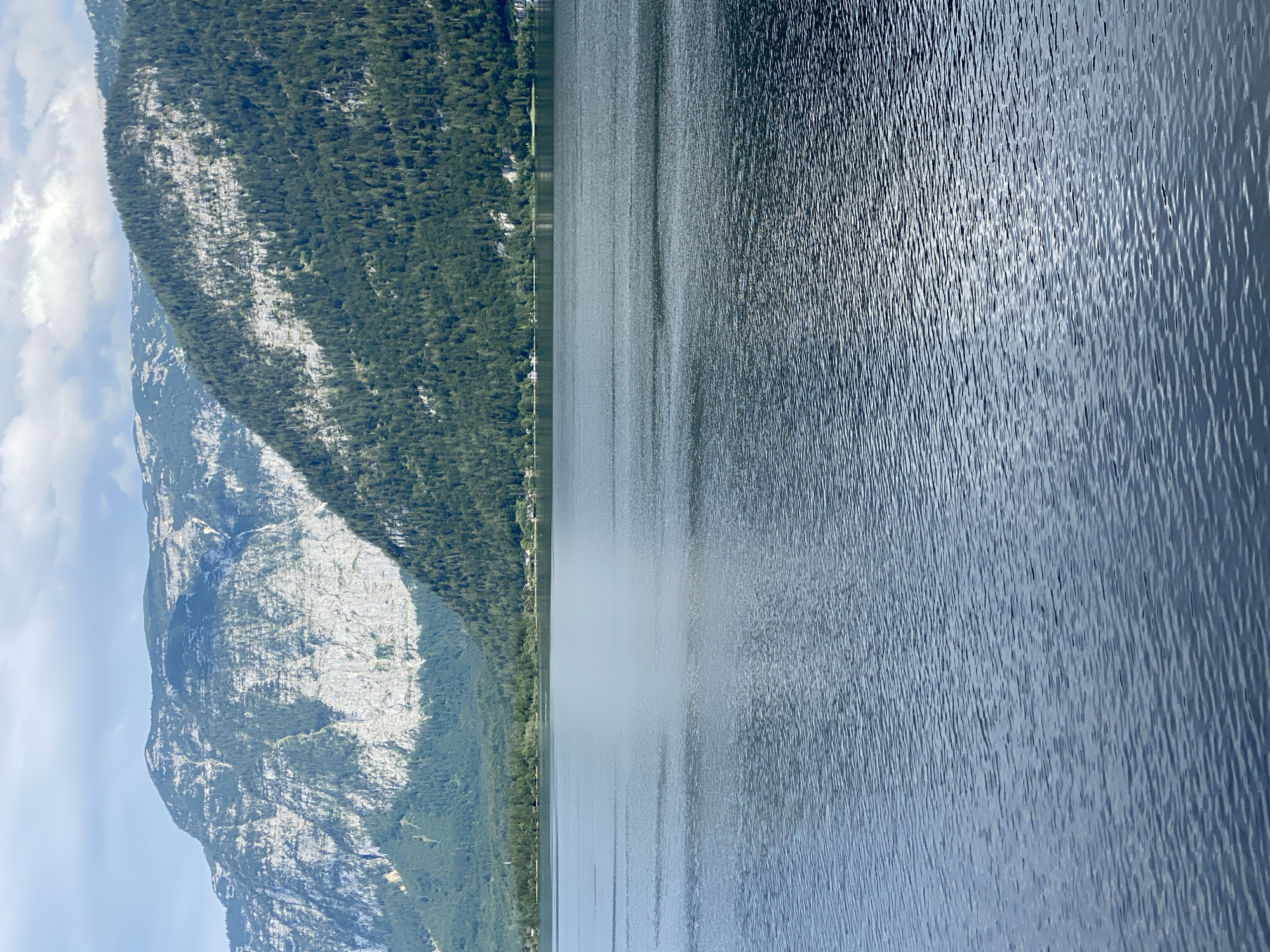 The image depicts a wide, still lake in the foreground, with gentle ripples disturbing its surface. In the background, a dense forest blankets the lower slopes of towering mountains. The mountains themselves are characterized by sheer rock faces interspersed with patches of vegetation. A thin strip of trees and some scattered buildings are visible along the shoreline, indicating a settlement. The sky above is partly cloudy, with patches of blue sky visible between the white clouds. The scene evokes a sense of natural beauty and tranquility, characteristic of the Austrian Alps. This view is likely from the town of Hallstatt, Austria, a UNESCO World Heritage site renowned for its picturesque setting on the Hallstätter See.