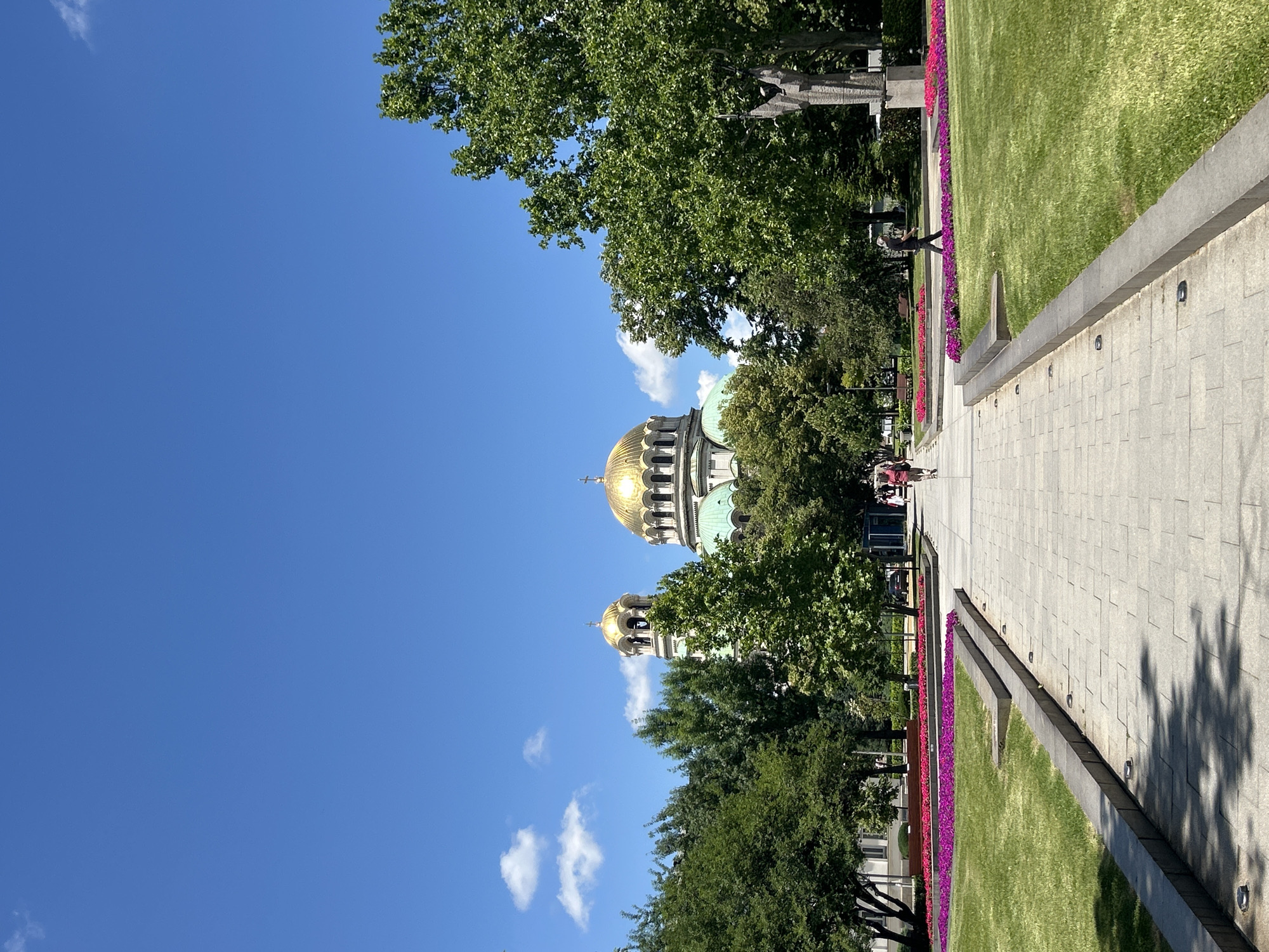 This is a vertical, outdoor, eye-level shot of a paved walkway leading towards a large, domed cathedral with golden domes, surrounded by green trees and manicured lawns. The sky is bright blue with a few scattered white clouds. In the foreground, the walkway is made of light gray rectangular stones. Flanking the walkway are raised, dark gray stone borders. Beyond the borders, there are large grassy areas with colorful flowerbeds in shades of pink and purple. Several people are visible: two individuals are walking away from the camera on the walkway in the middle distance, and one person is walking towards the right edge of the frame on the right side of the walkway. There is a statue on the right side of the image, partially obscured by trees. The cathedral, with its distinctive green and gold domes, is the prominent background subject, with trees partially obscuring its facade.