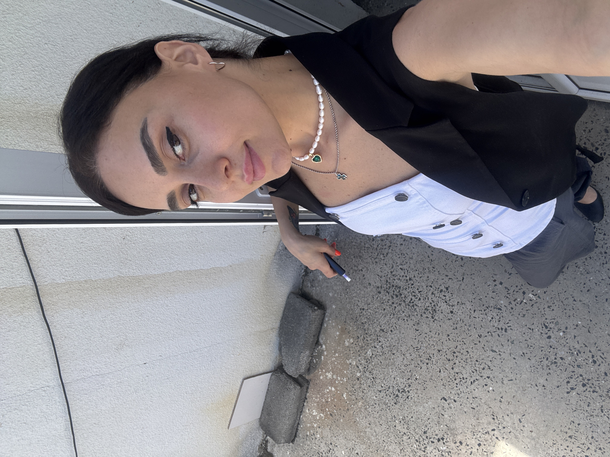 A young woman with dark hair and dark eyes is taking a selfie outdoors. She is wearing a white vest over a black blazer, dark gray pants, and black shoes. She has on a pearl necklace with a green heart pendant and a silver cross pendant. She is holding a dark blue object with a red tip in her right hand. Behind her is a rough, white wall and a concrete ground with some dark specks. There are two rectangular grey objects leaning against the wall, one with a white rectangular object partially covering it. The image appears to be taken in an outdoor urban setting, possibly an alleyway or a side street, with elements suggesting construction or renovation, such as the grey rectangular objects and the rough wall. The lighting indicates it is daytime with sunlight casting shadows. The environment suggests a location in Tbilisi, Georgia, due to the architectural style of the wall and the overall urban aesthetic.