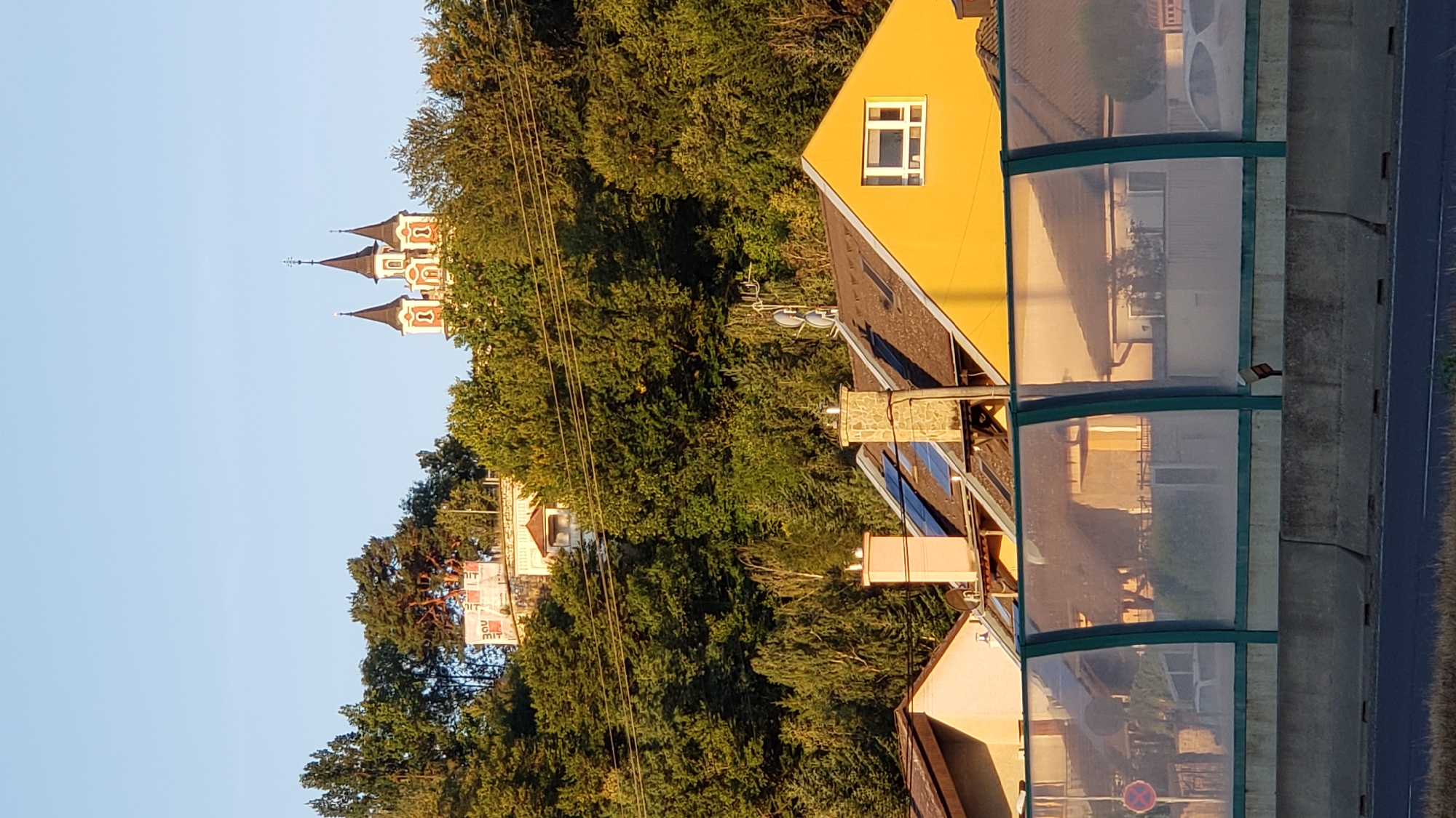 The image shows a view of a hill with buildings and trees in Prešov, Slovakia. At the top of the hill, a church with three spires is visible, surrounded by lush green trees. Below the church, other buildings and structures can be seen nestled into the hillside. In the foreground, a bright yellow house with a window is prominent, with a portion of a noise barrier and a concrete barrier in front of it. The scene is illuminated by sunlight, casting long shadows and creating a warm glow. The sky above is clear and blue.