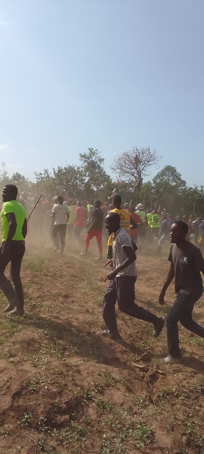 This outdoor image depicts a large group of people, predominantly young men, actively moving through a dusty, open area in Isukha ICHINA, Kenya. The scene is illuminated by bright daylight under a clear, pale blue sky, suggesting a mid-morning to late afternoon setting.
In the foreground, two men are running; one on the left in a bright neon green shirt, and another on the right in a dark t-shirt with white text, looking back and shouting. Another man in a patterned short-sleeved shirt and dark pants runs in the mid-foreground, also appearing animated. The ground is dry, reddish-brown earth with sparse patches of green vegetation, and a significant cloud of dust is kicked up by the movement of the crowd, obscuring parts of the background.
The mid-ground is filled with numerous individuals, many wearing brightly colored clothing such as red, yellow, and neon green shirts. Some are holding long sticks or poles. Their collective movement generates the prominent dust cloud. In the background, a line of green trees and bushes frames the area, with one distinctive, large, bare-branched tree standing out.
On a yellow high-visibility vest worn by an individual in the mid-ground, the word "ENERGY" is clearly visible, with another partial word that might be "KENRAY". The overall activity suggests a dynamic event, possibly a race, a procession, a sporting competition, or a spirited community gathering, characterized by high energy and lively interaction.