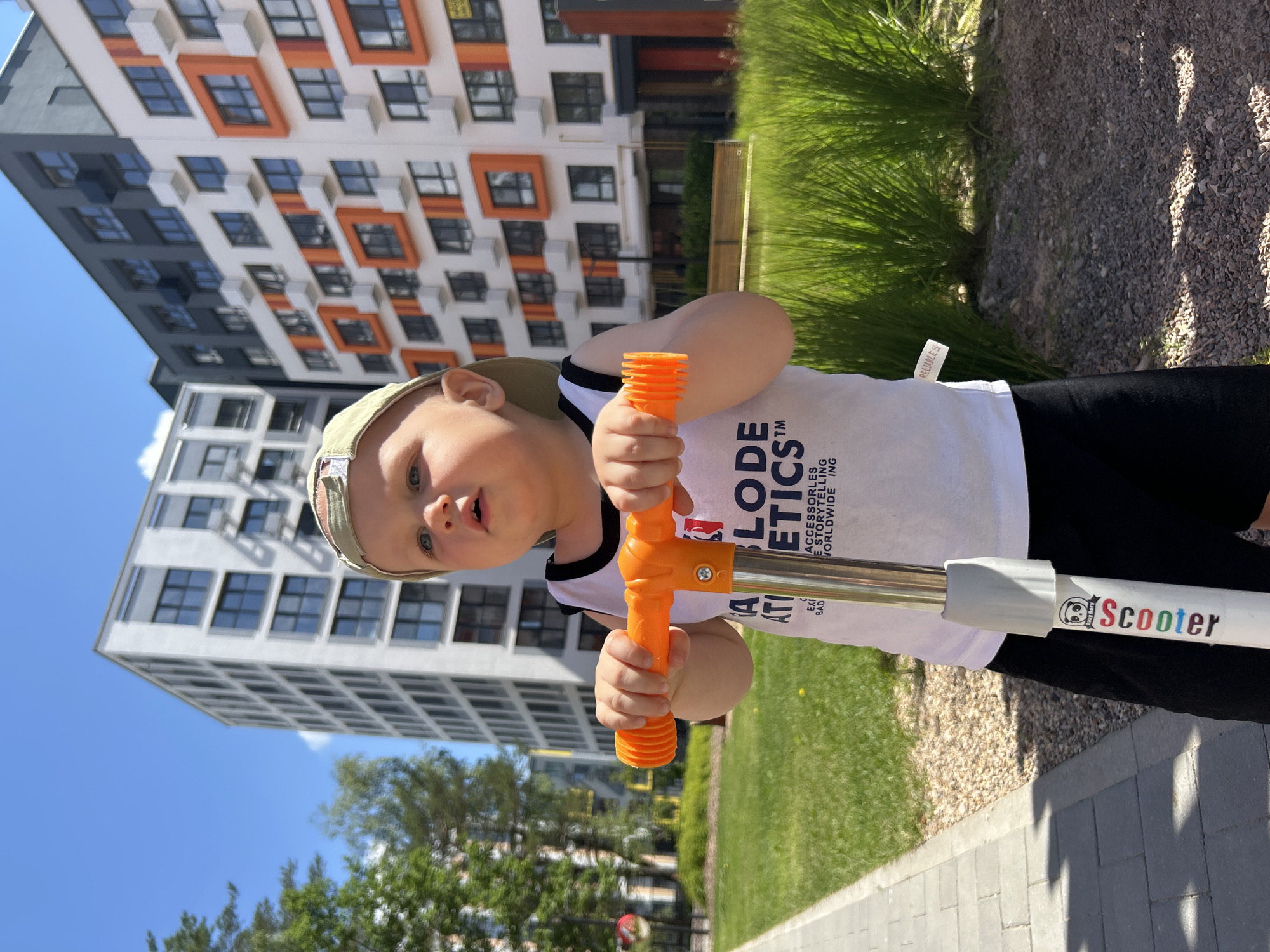 A young boy, wearing a light-colored baseball cap and a white t-shirt with black trim, is holding onto the handlebars of a scooter. The scooter has orange grips. He is looking towards the camera with an open mouth and wide eyes, suggesting surprise or excitement. The t-shirt has text printed on it, including "ABLODE ATICSTM" and "ACCESSORIES E STORYTELLING WORLDWIDE". A sticker on the scooter's pole reads "Baby Bor Dory Scooter" and "RELIABLE".
The boy is in an outdoor setting with a paved walkway in the foreground. Behind him, there is a lush green lawn and tall, thin grass. In the background, there are modern apartment buildings with white facades and orange-accented windows. The sky is a clear blue with some white clouds. The sunlight suggests it is daytime, likely a sunny day. The overall scene depicts a child enjoying time outdoors with his scooter in a residential area.