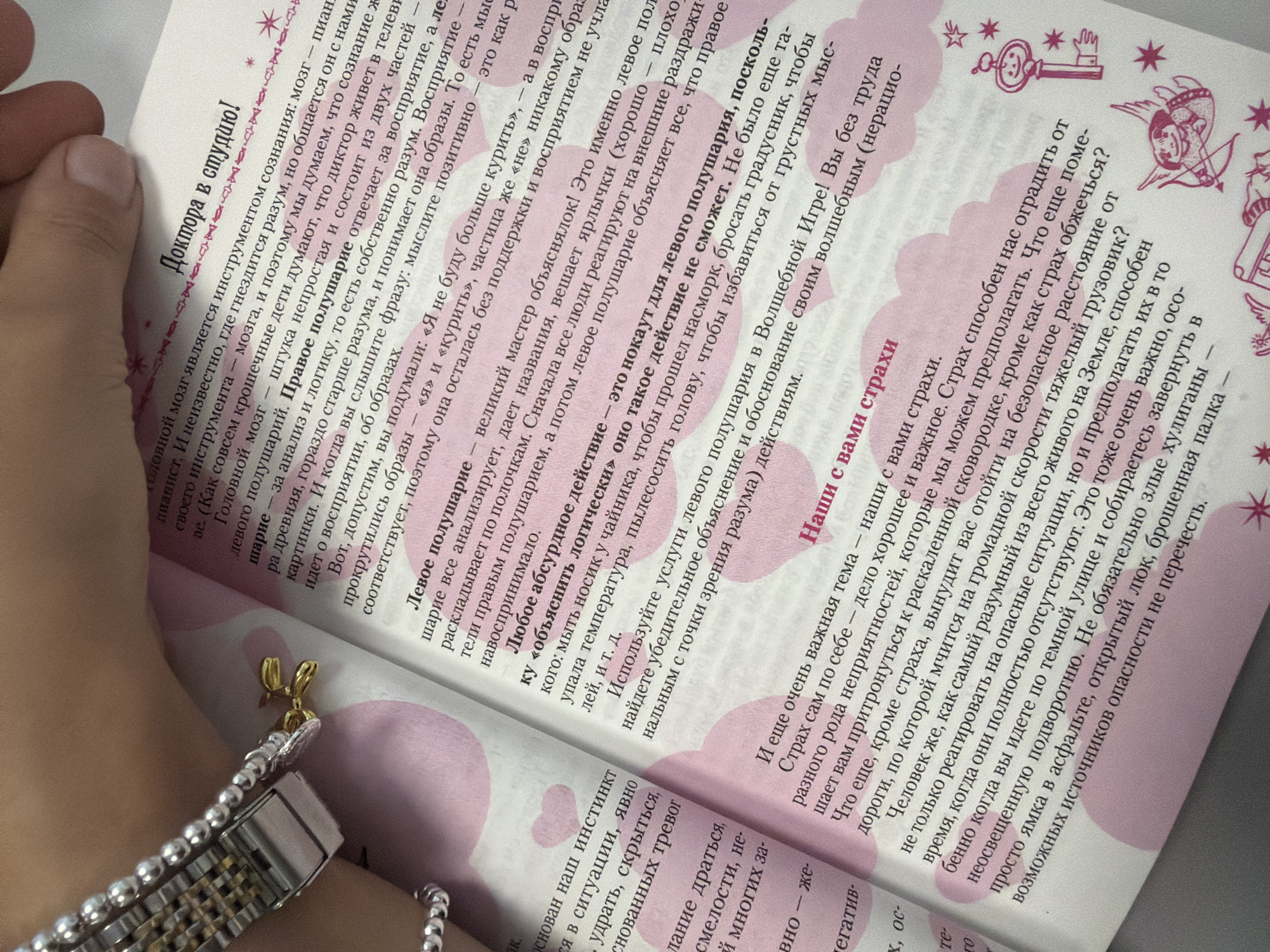 The image shows a close-up, overhead view of an open book with a pink and white patterned background. The book is open to a page with text in Russian. A person's hand, with fingernails painted pink, is visible on the right side of the image, holding the book open. A silver-toned watch with a beaded strap is on the left side of the image, also resting on the book. The book page features various headings and text in Russian. One heading in larger, bolder font reads "Доктора в студию!" (Doctors in the studio!). Another heading reads "Наши с вами страхи" (Our fears). The text discusses left and right brain hemispheres, cognitive functions, and fears. There are small decorative elements like stars and a key illustration on the page. The lighting suggests an indoor setting. There is no visible information in the image that places it in Homyel, Belarus, or any other specific city.