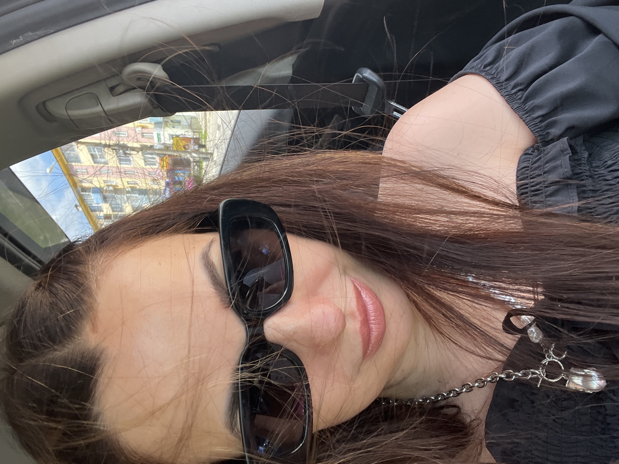 A medium close-up, rotated portrait shows a woman inside a car, looking towards the camera. Her head is tilted, and the image is rotated approximately 90 degrees counter-clockwise. She has long, dark brown hair, with numerous fine strands visible across her face, arms, and the car interior.
The woman is wearing large, black, rectangular sunglasses that cover a significant portion of her face. Her lips are a glossy pink, and she has well-defined eyebrows. She is wearing a black, off-shoulder top with ruffled or elasticized sleeves and a ruched neckline visible near her shoulder. A silver-colored chain necklace with a decorative charm or pendant is visible near her right shoulder and ear. Her skin tone is light.
The setting appears to be the interior of a car, indicated by the beige-colored headliner, a sun visor mechanism, and a seatbelt anchor visible in the background. Through the windshield, urban buildings with multiple windows are visible, suggesting a daytime, possibly overcast, environment. The buildings outside appear multi-storied and have varying colors, including pink, yellow, and possibly blue.
The woman appears to be posing for a photograph, likely a selfie, given the direct gaze into the lens. No specific activity beyond posing is evident. There is no visible text in the image.