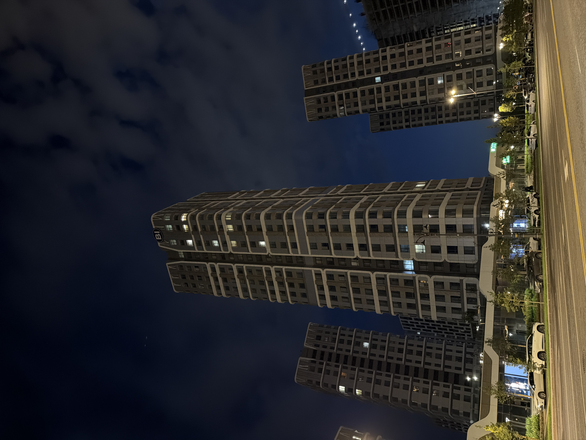 This image shows a night scene in Astana, Kazakhstan, featuring modern high-rise buildings and a street-level commercial area. The sky is dark blue with scattered clouds, and some lights are visible on the horizon, possibly from other buildings or a bridge.
The focal point is a cluster of several tall, contemporary apartment buildings with a uniform facade characterized by alternating light and dark rectangular windows and decorative vertical bands. The building in the foreground on the right has a large "BI" logo at its peak. Some windows are illuminated, indicating occupancy. To the left, three other similar buildings are visible, two of which are partially obscured by the foreground building. Further to the right, an unfinished or under-construction building is present, with exposed structural elements and a string of lights.
At the base of the buildings, a modern commercial strip is visible with a curved roofline. Several trees are planted along the sidewalk, some illuminated by streetlights. A street with cars parked along the side runs in front of the commercial area. At least five cars are visible on the street, including white sedans and a darker colored SUV. The street is paved with asphalt and marked with yellow lines. The overall impression is one of a modern urban development at night.