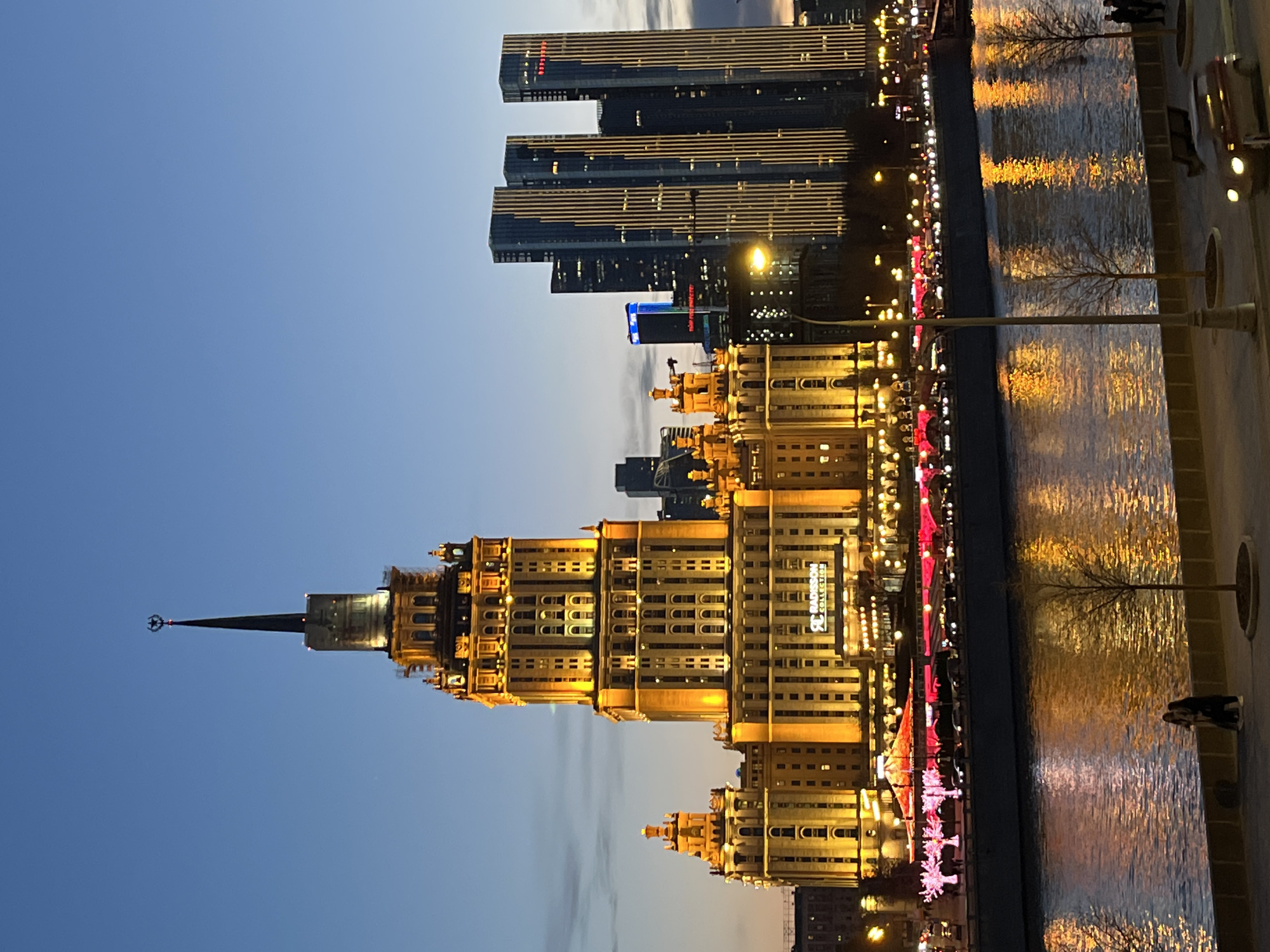The image depicts a night scene in Moscow, Russia, featuring the Kotelnicheskaya Embankment Building, one of the Seven Sisters skyscrapers, prominently illuminated in golden light. Tall modern skyscrapers are visible in the background. A river reflects the lights of the city. A pedestrian area with lamp posts runs along the right side of the image. Bare trees line the waterfront.