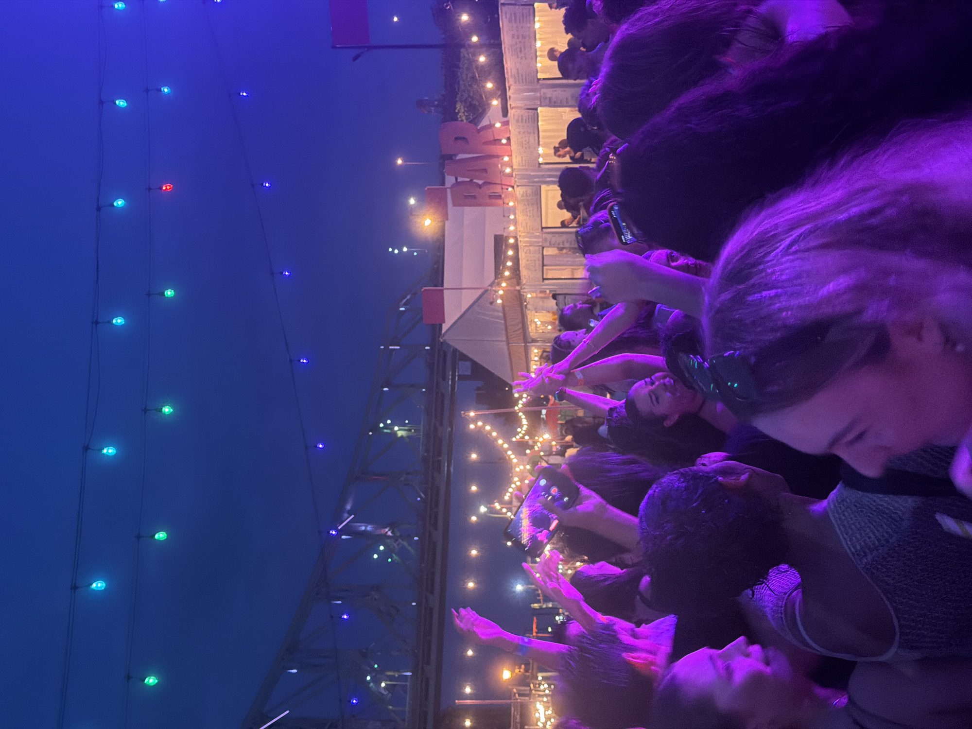 The image depicts a nighttime outdoor event with a large crowd of people. The atmosphere is festive, with many individuals raising their hands. String lights, in blue and green, are visible overhead. In the background, a large bridge structure is illuminated, and there is a building with a prominent "BAR" sign. The overall lighting is purple, suggesting a concert or party setting. The presence of a bar and the gathering of many people indicate a public venue. Based on the bridge and the overall urban aesthetic, the location is likely an outdoor event space within the city of Montreal, Canada.