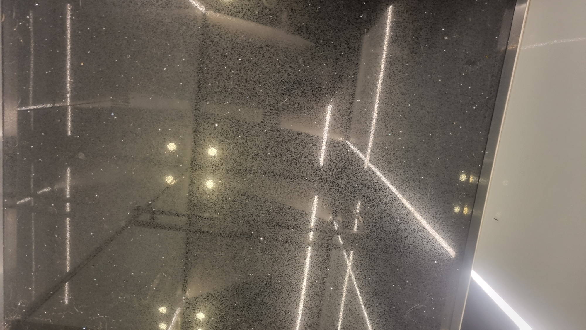 This media file is a close-up, high-angle shot of a dark, speckled surface, likely a countertop or a wall panel with a granite or terrazzo-like finish. The surface is highly reflective, showing multiple bright, linear light reflections. These lights appear to be recessed or integrated into the ceiling or surrounding structure, creating long, sharp white lines that traverse the frame diagonally and vertically. The speckled texture of the surface is visible, with numerous small white and light-colored particles embedded in the dark material. There are also some scattered, blurred circular reflections that appear to be from overhead light fixtures. A thin, silver-colored metallic trim runs vertically along the right edge of the dark surface, separating it from a lighter, smooth, off-white surface on the right. The off-white surface also has some faint reflections and what appears to be a small dark insect or speck on it. The scene is well-lit, with strong contrasts between the dark surface and the bright light reflections. There are no people, animals, or clear indications of weather or time of day beyond artificial lighting. The location cues are minimal, suggesting an indoor, modern, and possibly commercial or residential setting.