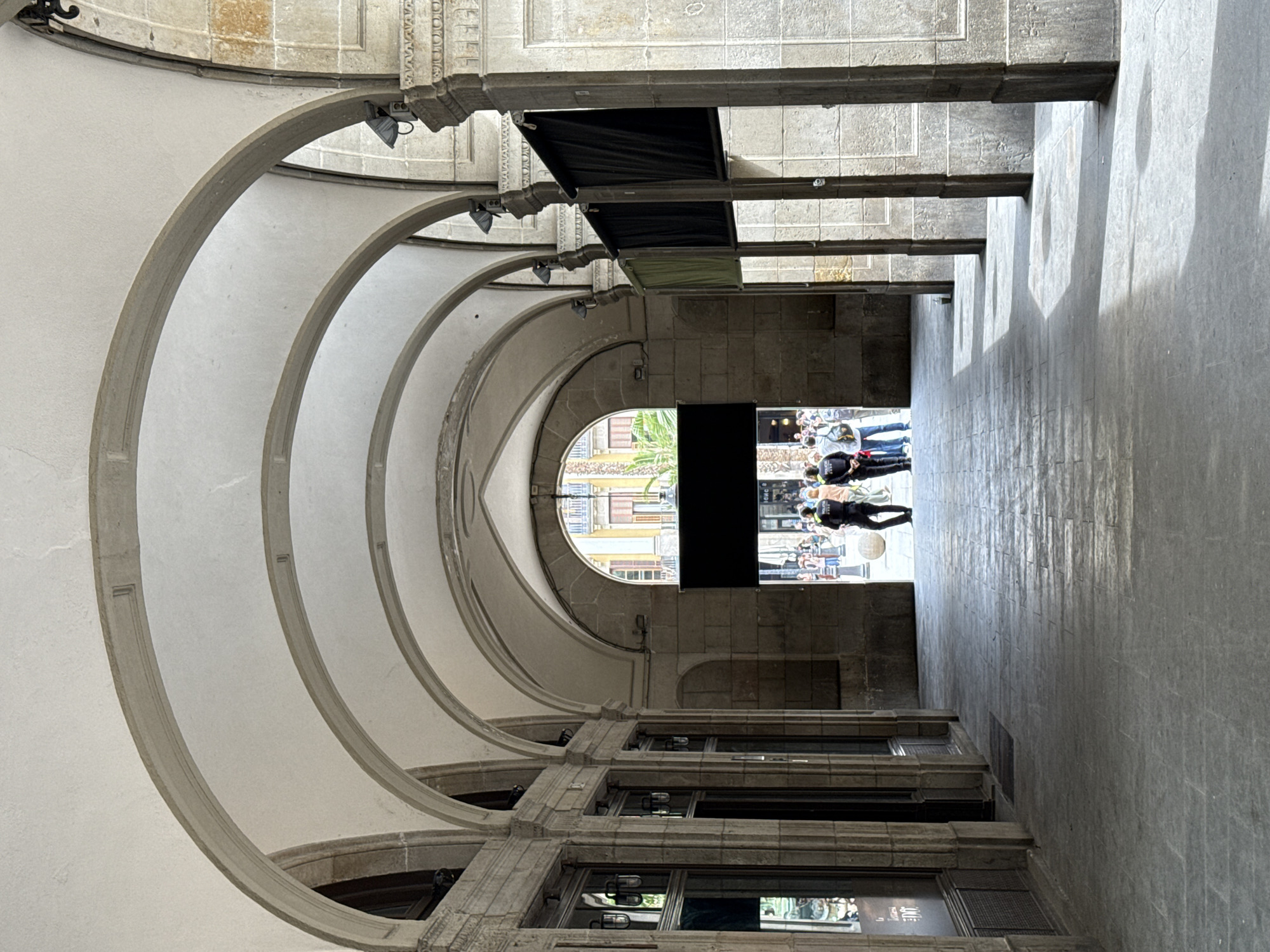 This photo depicts a view down a narrow corridor with a series of arched ceilings. The architecture appears old, with stone structures and repeating archways. There are people in the distance at the end of the corridor. Black awnings hang from the ceilings of the corridor. The photo was taken from Barcelona, Spain.