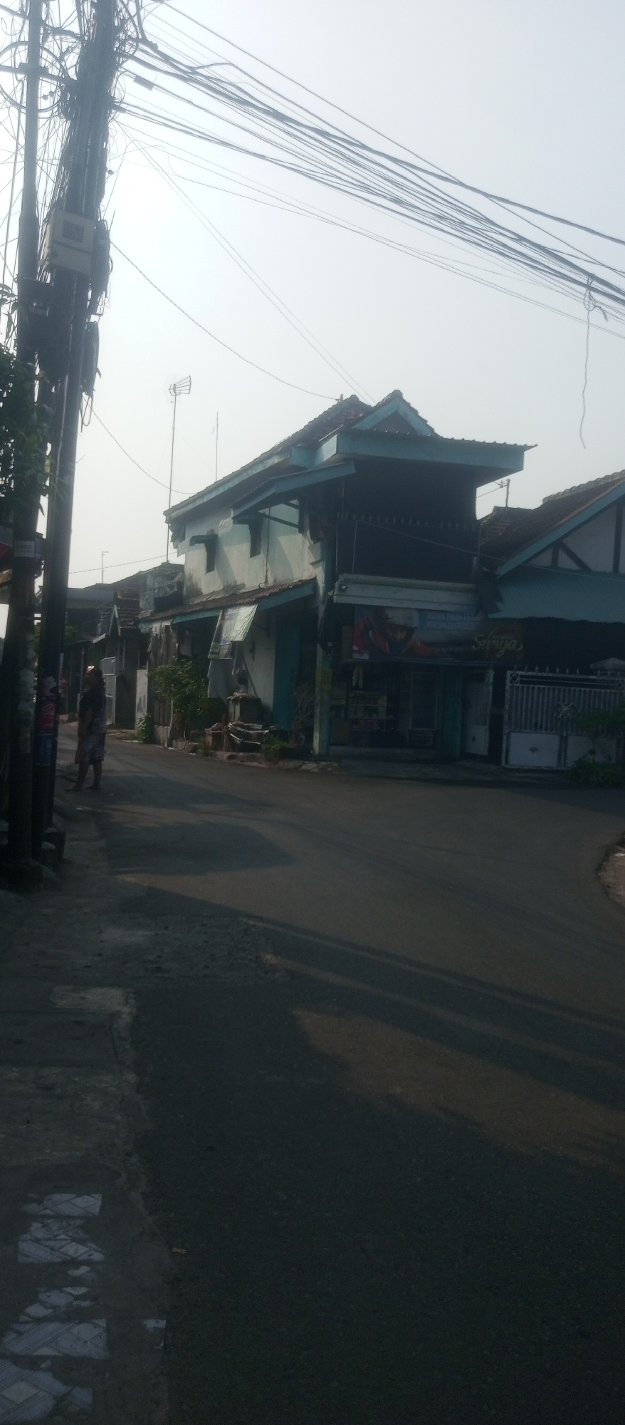 A residential street scene in Kediri City, Indonesia, bathed in the hazy morning light. A person stands on the sidewalk near a utility pole laden with a dense network of electrical wires. The street itself is paved and mostly empty, with shadows stretching across it. On the right side of the street, a row of modest houses and shops with faded blue trim line the road. One shop, identified by a sign reading "Surya," appears to be open. The architecture suggests a common urban residential style in the region. The overall impression is one of a quiet, ordinary morning in a built-up area.