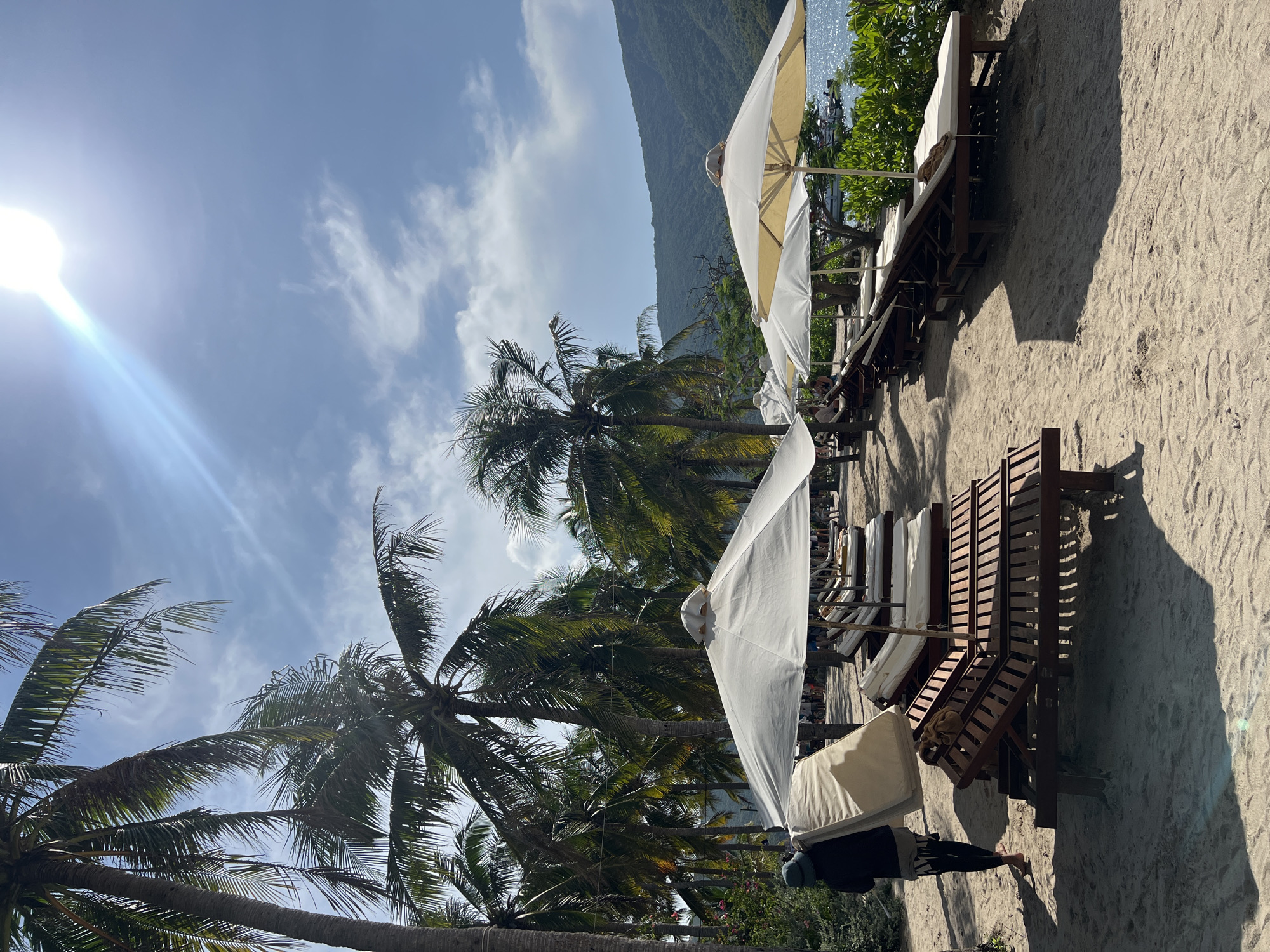 The scene depicts a beachfront in Nha Trang Ward, Vietnam. Numerous lounge chairs with white cushions and umbrellas line the sandy beach, casting shadows. Palm trees are interspersed between the umbrellas. A mountain is in the background, partially obscured by clouds. The sky is bright blue with scattered clouds. A person, wearing a hat and dark clothing, stands near one of the lounge chairs.