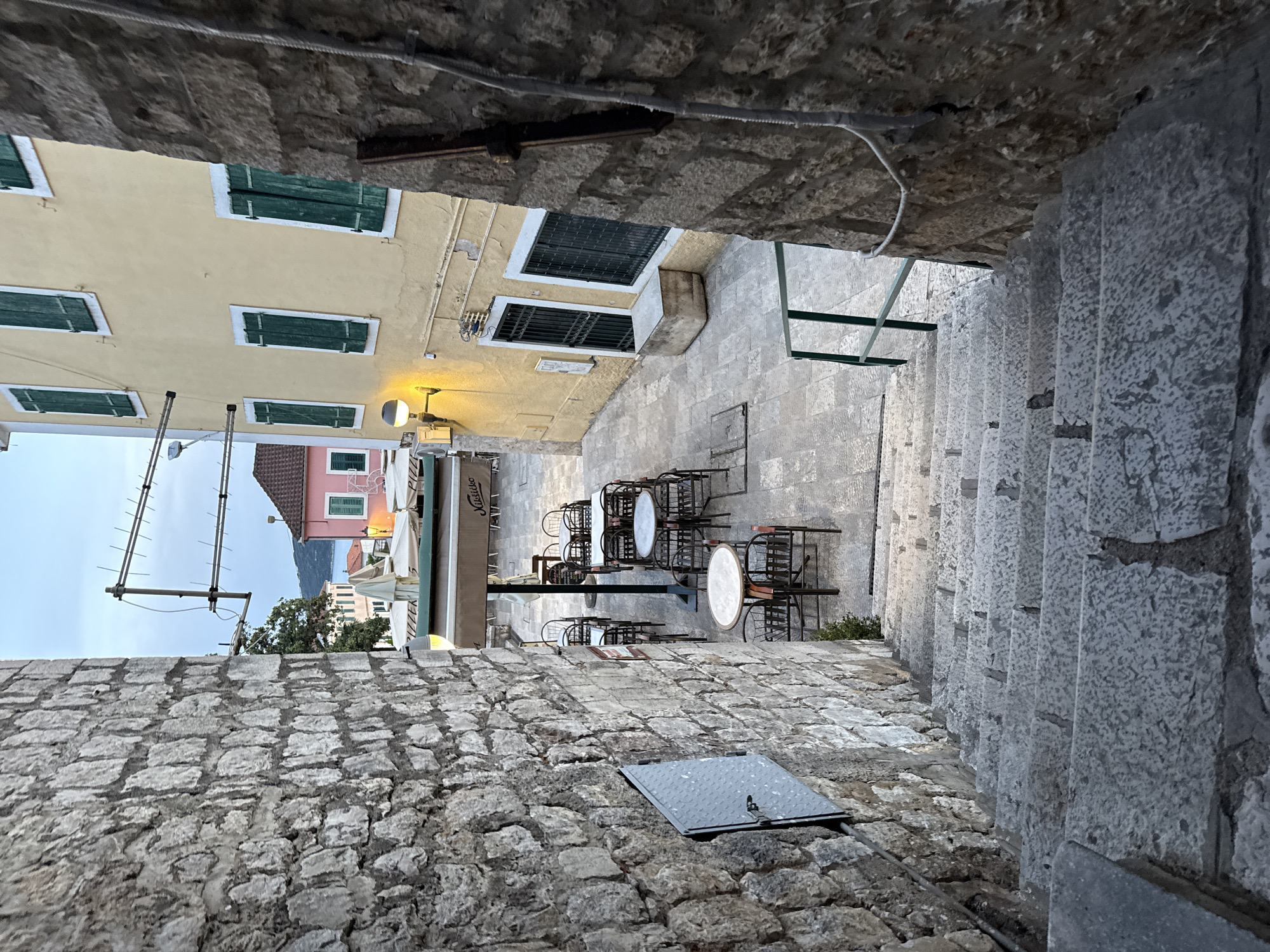 An eye-level, upward-looking view from the bottom of a stone staircase in Herceg Novi, Montenegro, reveals a narrow alleyway framed by weathered stone walls. The staircase, composed of irregular gray stone steps, ascends to the right. The alleyway opens to a small, stone-paved plaza with outdoor cafe seating, including round tables and chairs. Buildings with yellow and pink facades and green shutters line the plaza. An antenna is visible extending above the buildings. Cafe umbrellas and signage are present. A manhole cover is embedded in the stone wall to the left. Overhead, exposed wires and the undersides of the stone structures form a partial ceiling. No people are visible.