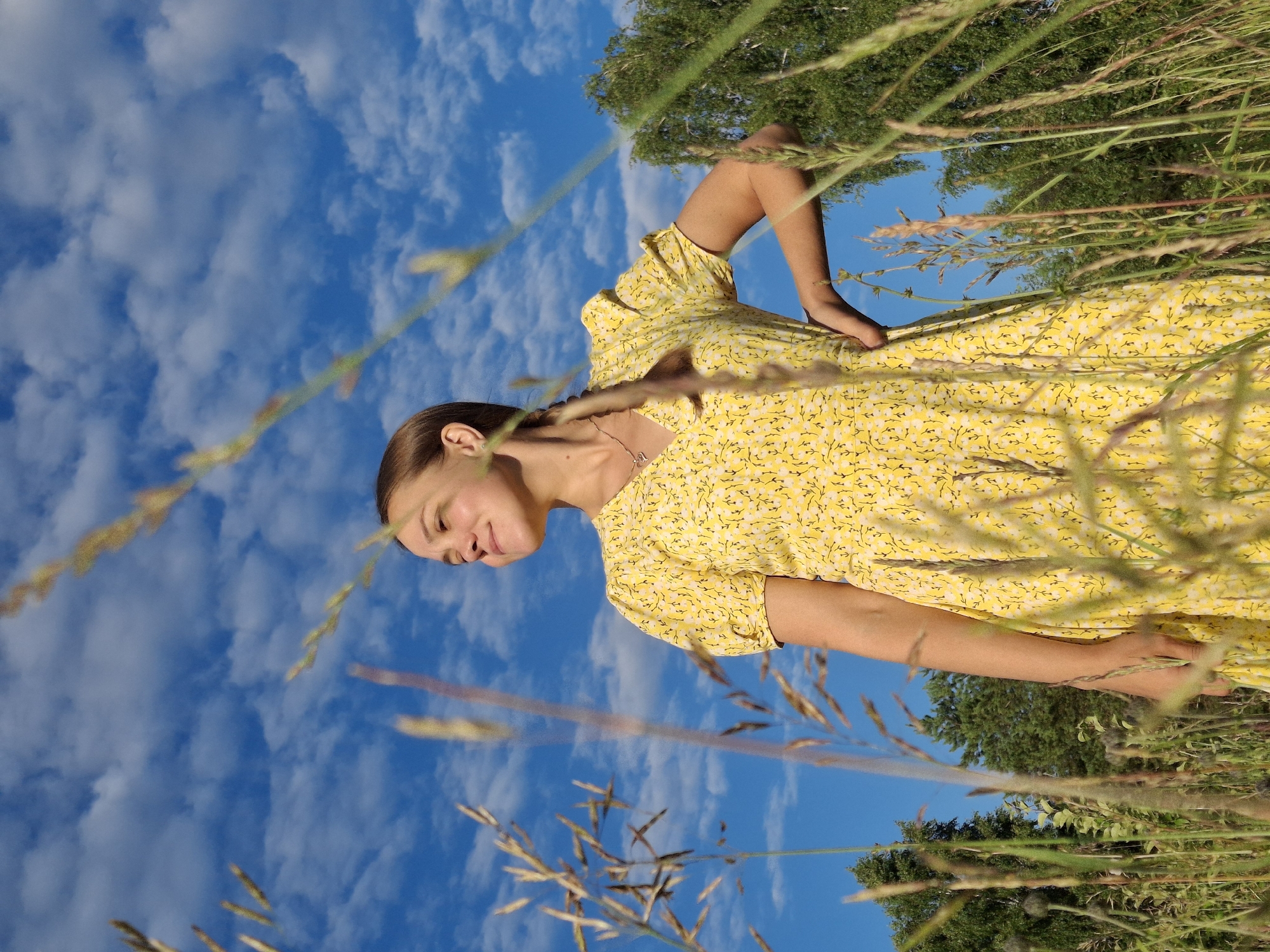A young woman, likely a teenager, is the subject of this photo, taken outdoors in Chaikovsky, Russia. She is wearing a bright yellow dress with a small floral pattern. The dress has short sleeves and a cinched waist. Her hair is pulled back in a braid. She is looking down and to the side with a gentle smile, suggesting a relaxed and content mood. The photo is taken from a low angle, with the blue sky and white clouds filling most of the background. Tall, wispy blades of grass are in the foreground, some in sharp focus and others blurred, creating a sense of depth and framing the subject. A large green tree is visible in the upper right portion of the image. The lighting suggests it is daytime, possibly late afternoon given the warm quality of the light. The overall impression is one of a peaceful summer day in a natural setting. There is no visible text in the image.